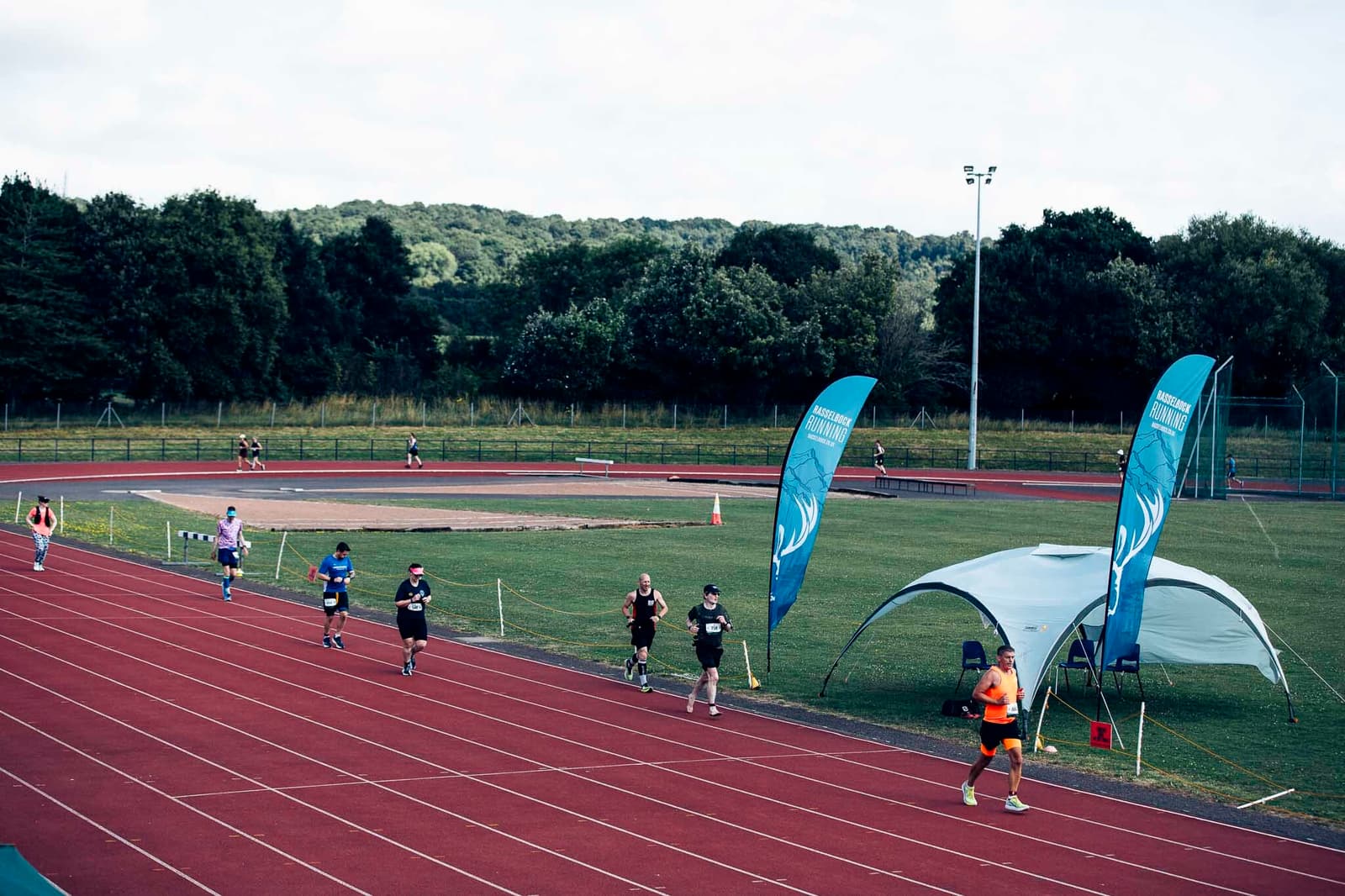 Runners participating in a race on a red track, flanked by teal banners. A tent is set up on the grass beside the track. Trees and a hill are visible in the background under a cloudy sky.