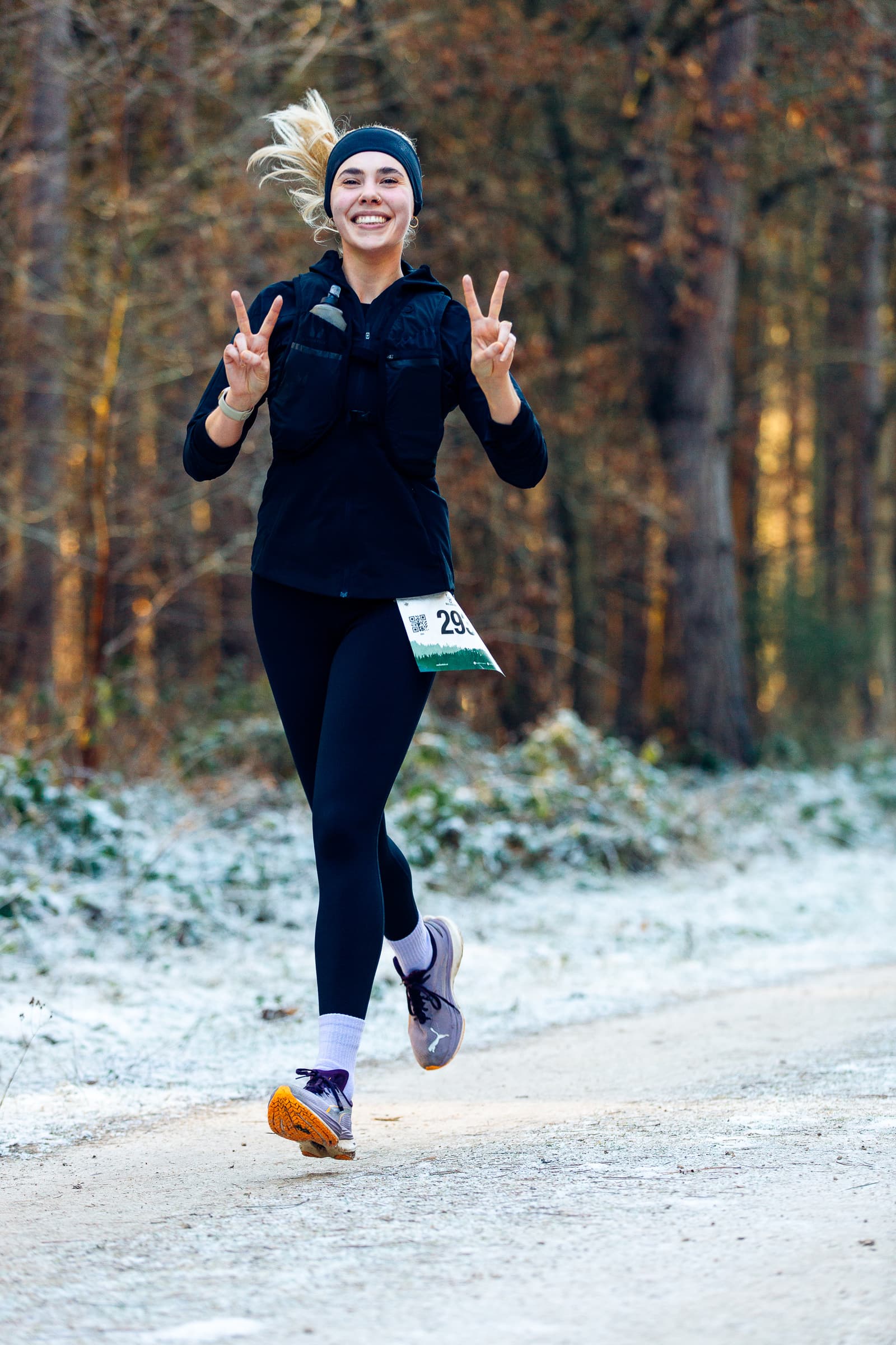 A woman jogs on a frosty woodland path, smiling and flashing peace signs with both hands. She's wearing a black outfit, headband, and bib number 29. Tall trees and a light dusting of snow line the path.