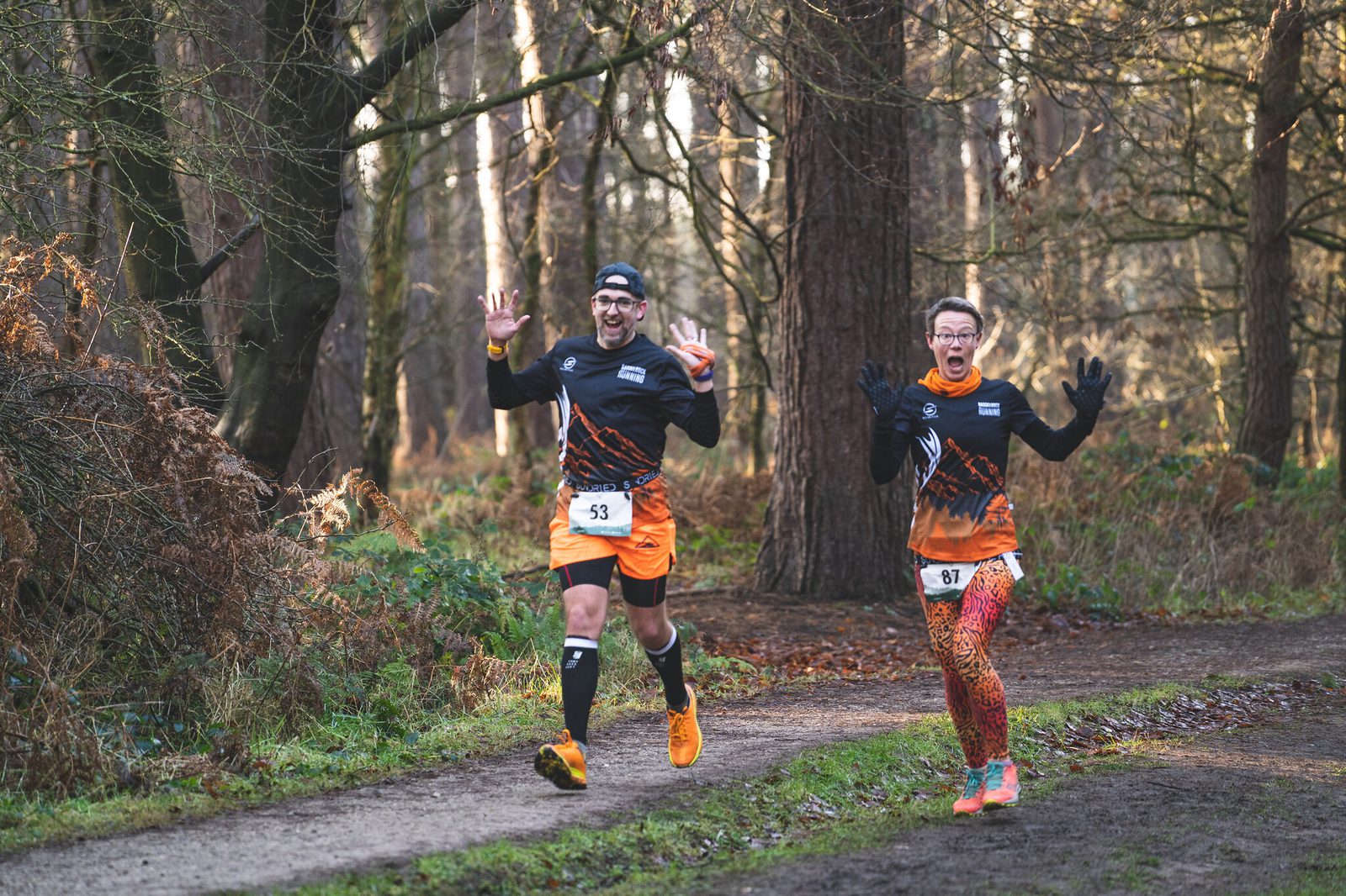 Two runners in vibrant athletic gear jog through a wooded trail, holding up their hands with excited expressions. They have race bibs numbered 53 and 87. The background features tall trees and foliage, suggesting it's a fun run or race in a forest setting.