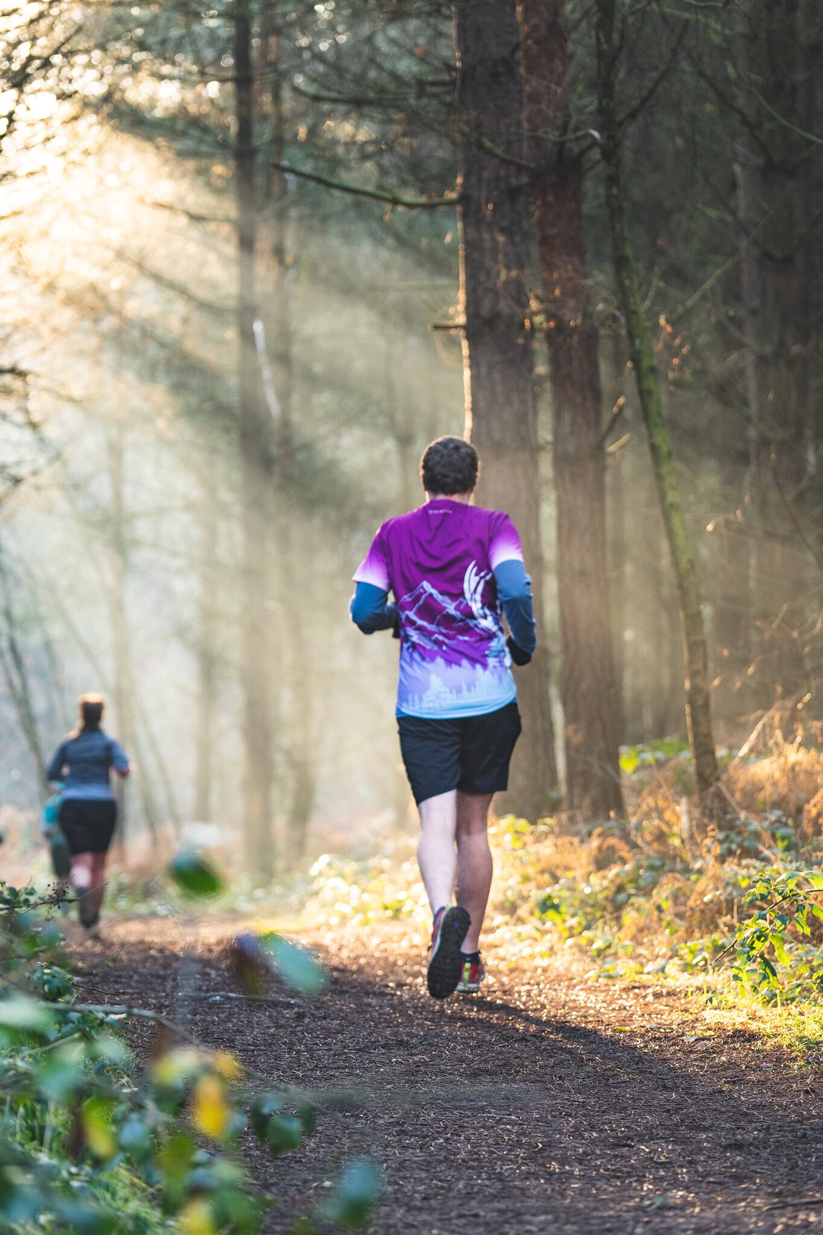 Two people running on a forest trail with sunlight streaming through the trees. One runner in the foreground wearing a purple and blue shirt and black shorts; the other is further down the trail, partially obscured by the trees. The scene is tranquil and sunlit.