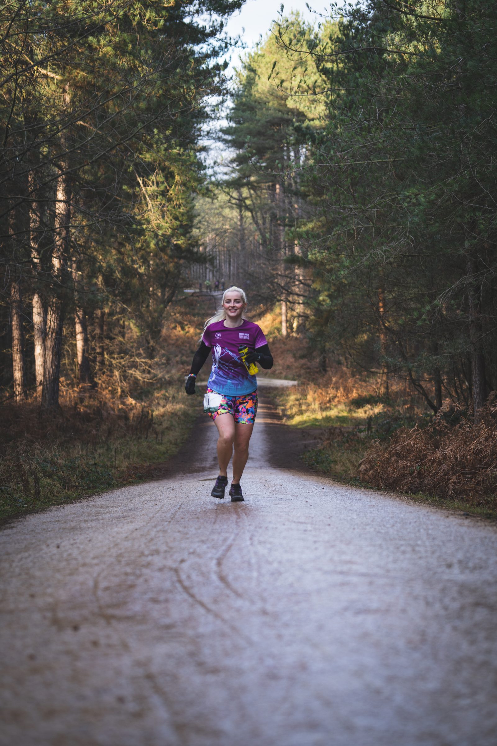 A person wearing colorful running shorts and a purple shirt runs along a gravel path through a forest. The sun filters through the trees, casting dappled light onto the pathway. The runner appears joyful and immersed in the natural surroundings.