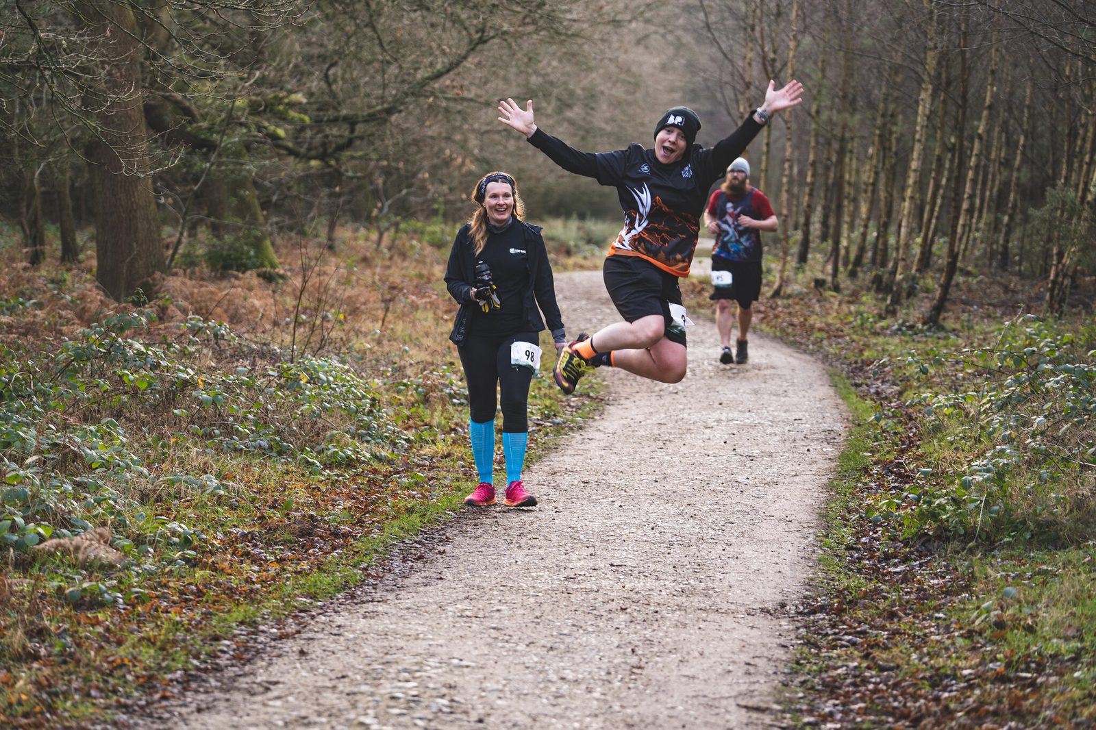 A man in a black outfit and beanie jumps excitedly mid-air on a forest trail, raising his hands high. Beside him, a smiling woman in a black jacket and bright blue leggings jogs. Another runner follows in the distance. Trees and foliage surround the path.