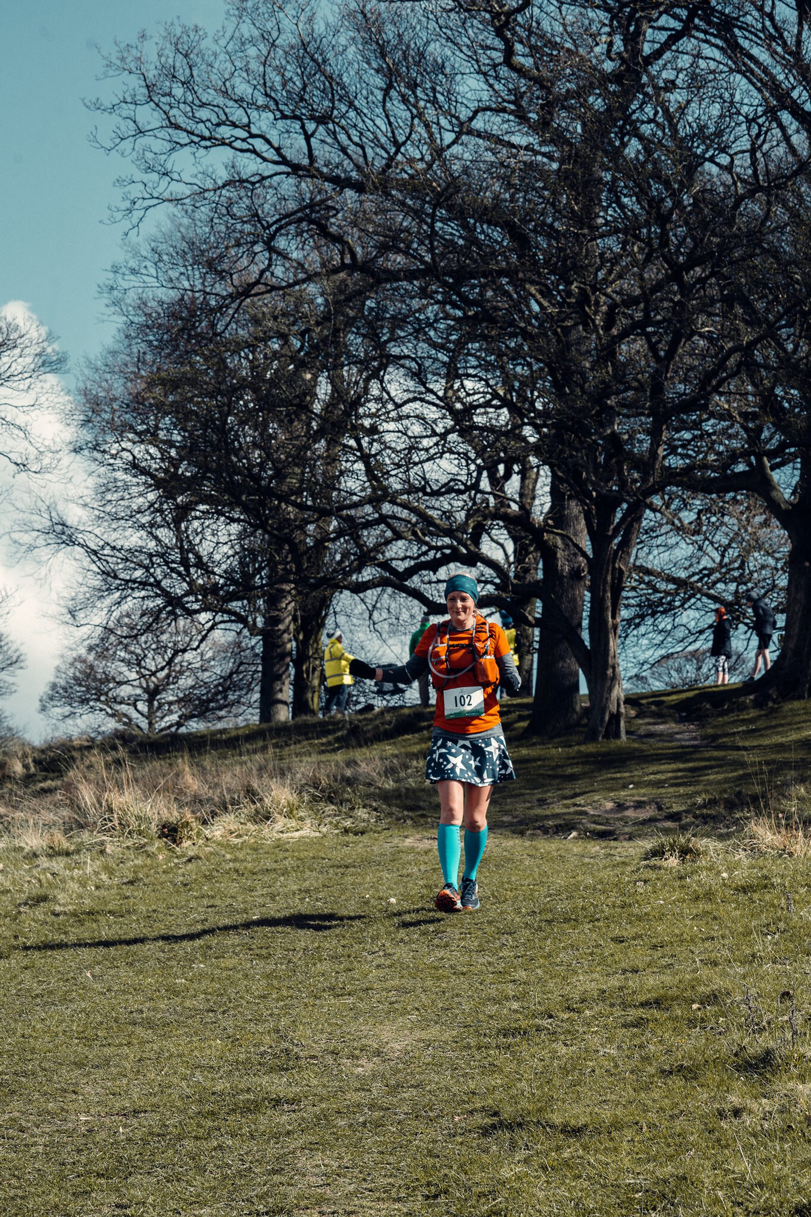 A runner wearing a red race bib numbered 102 runs down a grassy hill. The person is dressed in colorful gear, including a turquoise headband, orange shirt, and blue socks. Leafless trees and other runners can be seen in the background under a partly cloudy sky.