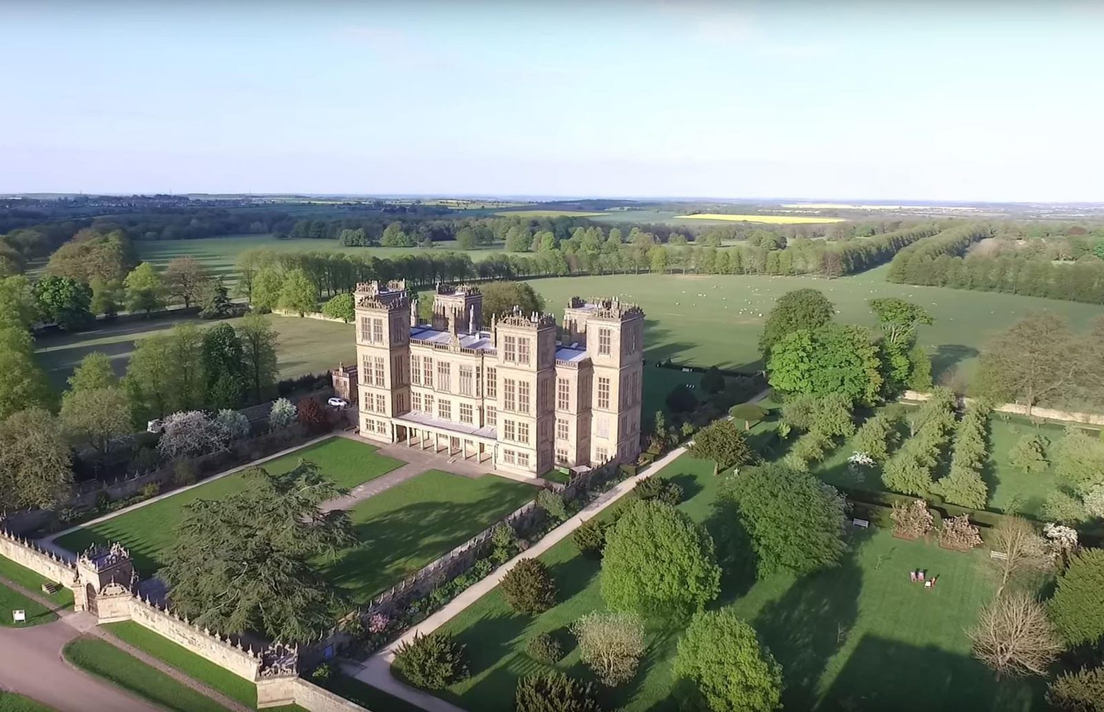 An aerial view of a grand, historic mansion surrounded by vast green lawns and trees. The large building has numerous windows and ornate architectural details. A long driveway leads to the entrance, and the estate is set amidst a scenic, expansive landscape.