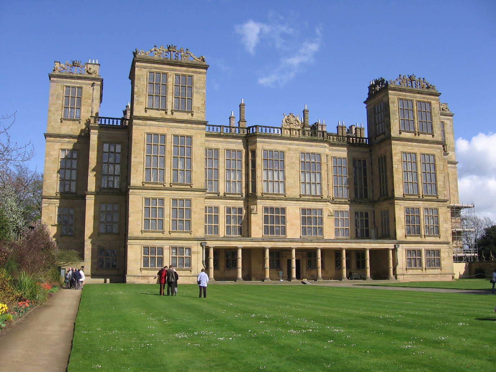 A grand, symmetrical Elizabethan mansion with large windows and ornate decorations stands against a clear blue sky. The extensive, well-manicured lawn in front has a few people walking and admiring the architecture. Colorful flowers line a pathway to the left.