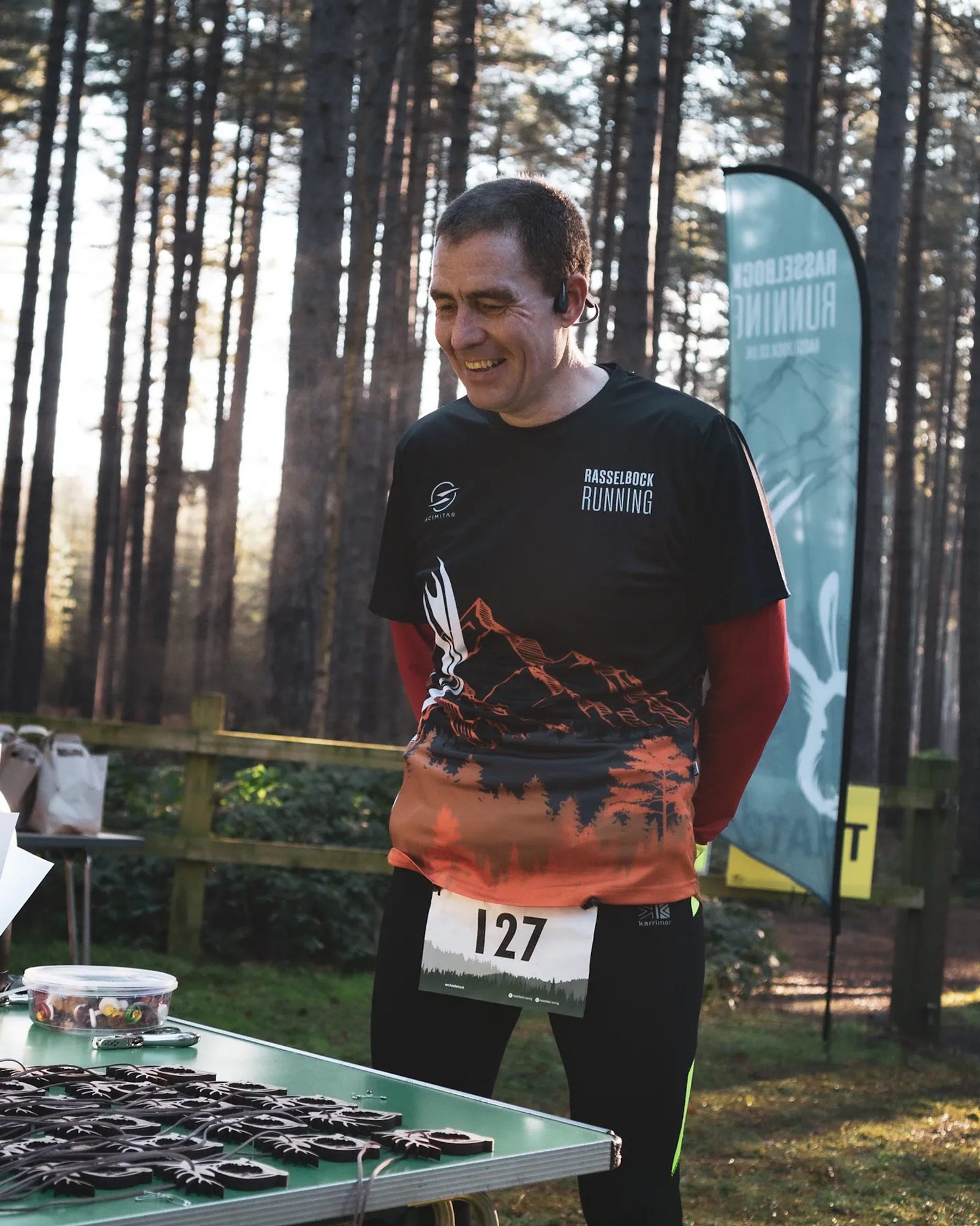 A person wearing a "Rasselbock Running" shirt and bib number 127 smiles while standing beside a table displaying medals in a forested area. In the background, trees and a green "Rasselbock Running" banner are visible.