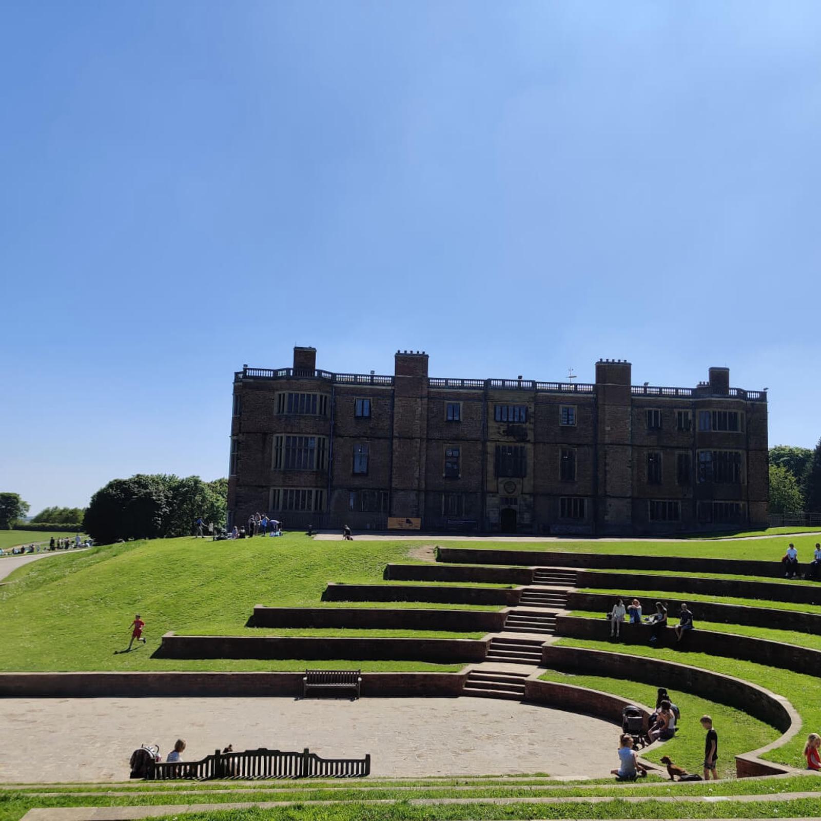 A historical brick building with large windows stands on a sunny day. In the foreground, people relax on tiered green grass steps and benches in front of the building. The sky is clear and blue.