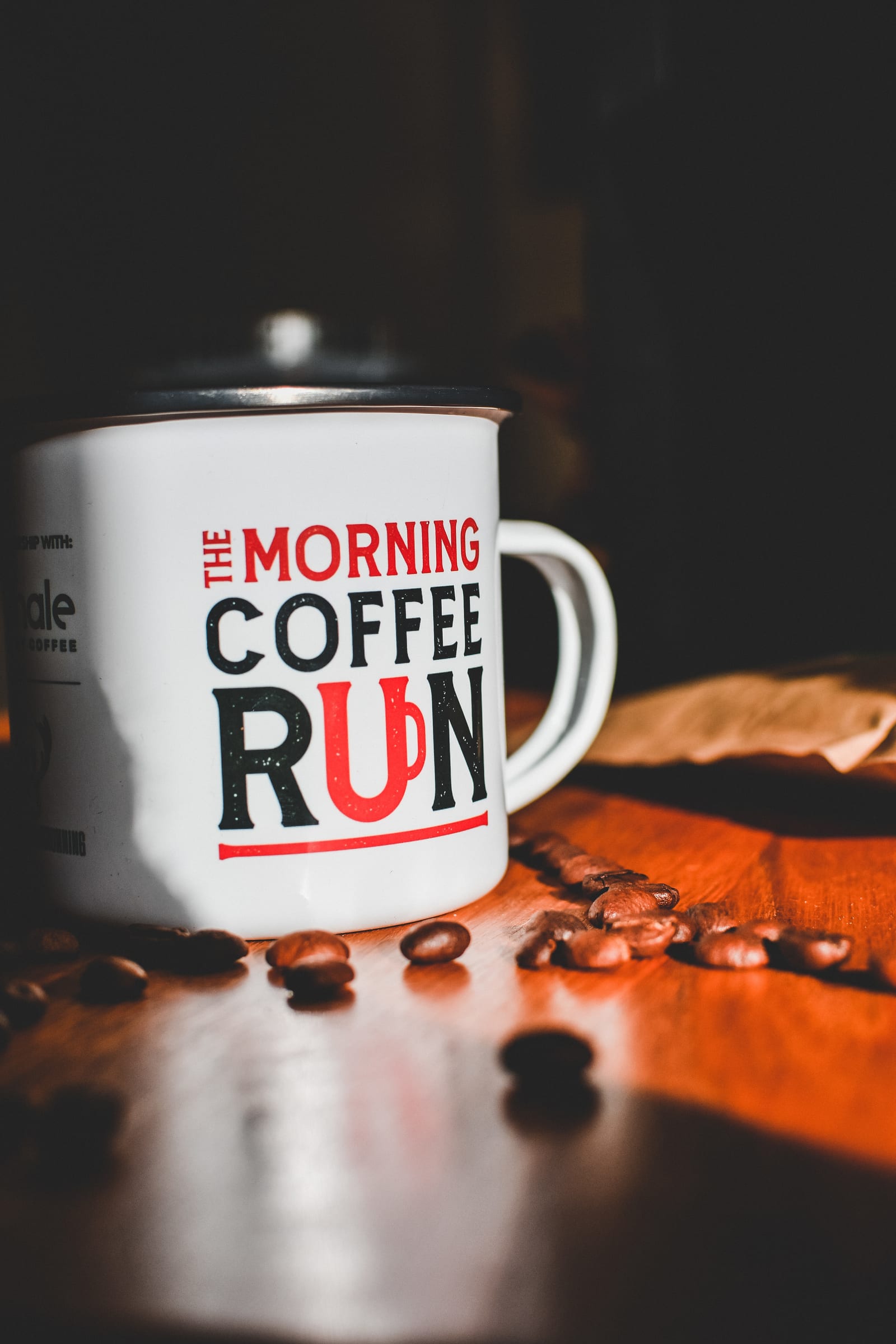 A white coffee mug with the words "THE MORNING COFFEE RUN" printed in bold red and black letters sits on a wooden surface surrounded by scattered coffee beans. Sunlight casts shadows on the mug and the beans, creating a warm and inviting atmosphere.
