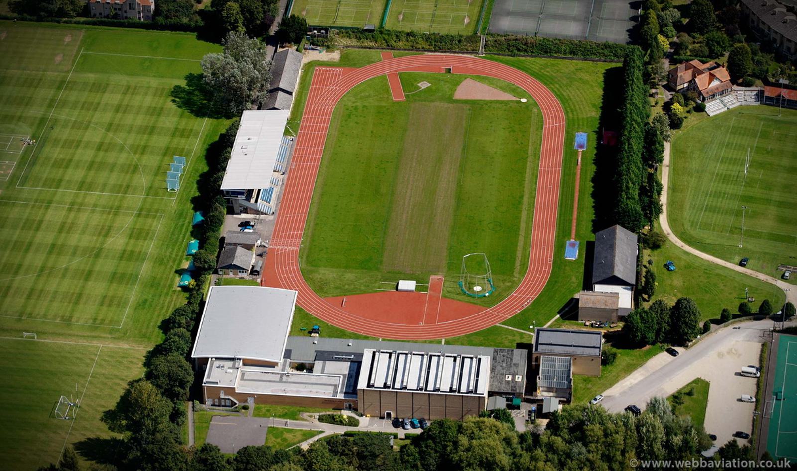 An aerial view of an athletics track with a red running surface surrounding a green sports field. Nearby, there are multiple buildings, additional sports fields, and trees surrounding the area. Tennis courts are visible in the upper part of the image.