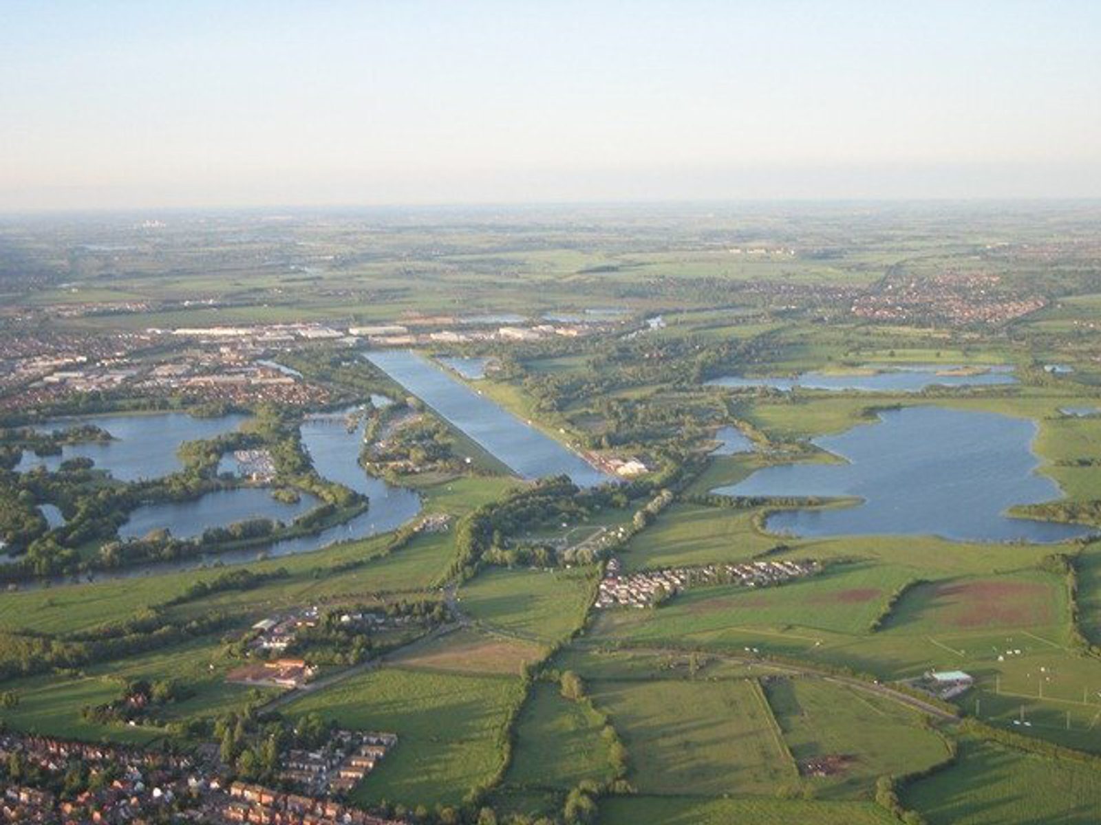 Aerial view of a landscape featuring multiple lakes, lush green fields, and a small residential area. A long, straight canal cuts through the center, flanked by trees and residential clusters. The sky is clear, suggesting a bright, sunny day.