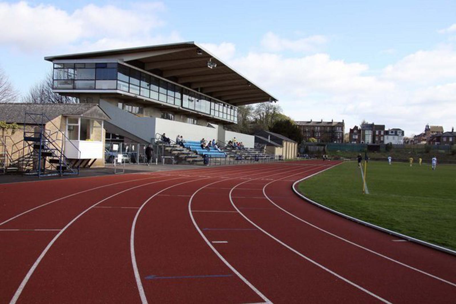 A red running track curves around a grassy field with a game in progress. In the background, there is a modern, multi-tiered spectators' stand with a roof, seating, and a press box. Further back, houses and buildings are visible under a partly cloudy sky.