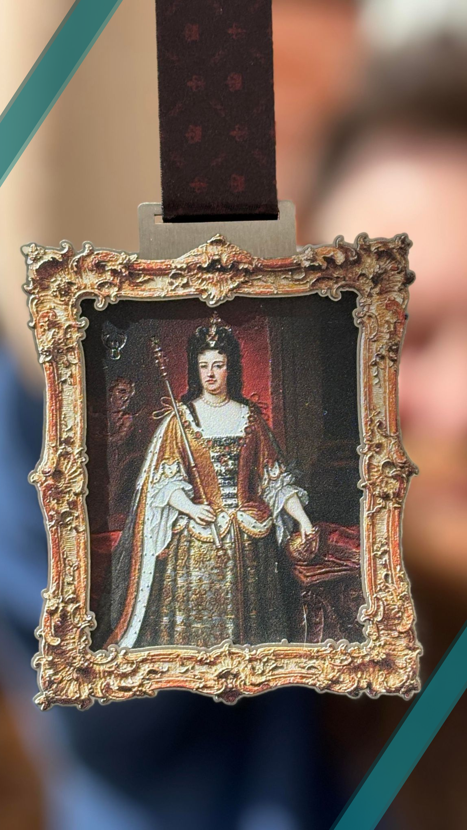 A decorative pendant featuring a portrait of a historical woman in regal attire, holding a scepter. The ornate frame has intricate golden details, and the background appears blurred.