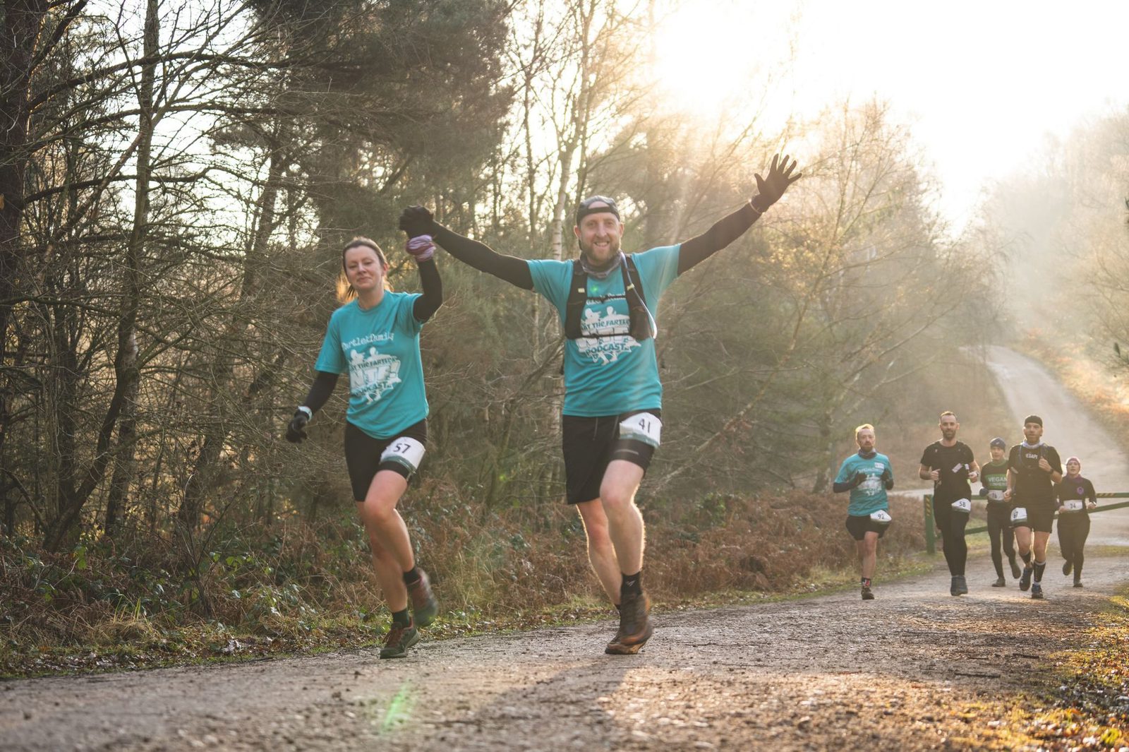 Two runners in turquoise shirts celebrate while running a race on a forested trail in the morning sunlight. They hold hands and raise their arms in joy. Other runners are seen behind them on the same path. The scene is bright and filled with energy.