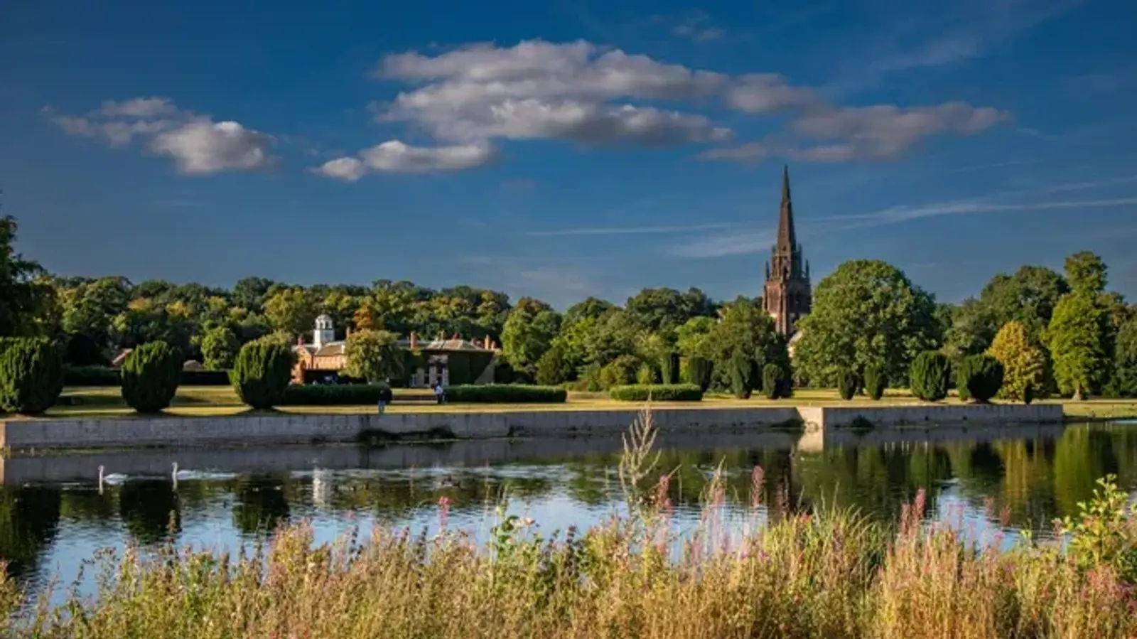 A scenic view of a park with a lake in the foreground, swans gliding on the water. In the background, there is lush greenery and a tall, pointed church spire against a blue sky with scattered clouds.