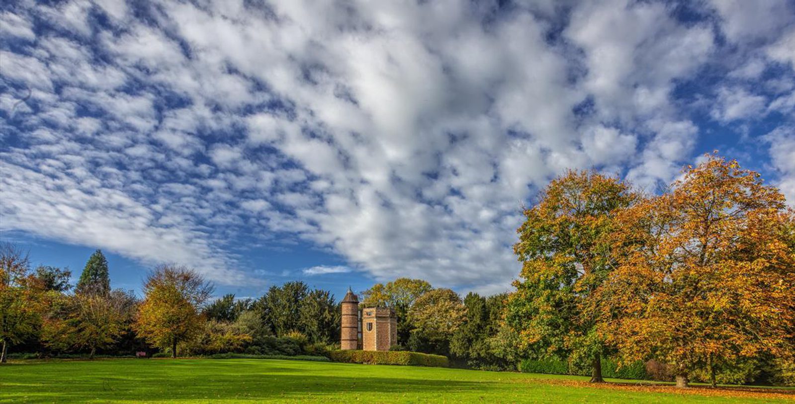 A picturesque landscape showcasing a well-maintained green lawn leading to a historic brick tower partially hidden by dense trees. The scene is under a dramatic cloudy sky, with autumn leaves on trees adding vibrant colors to the image.