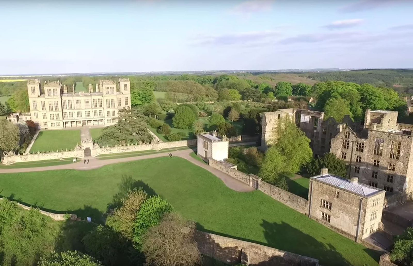 Aerial view of a historic estate featuring a grand manor with numerous windows, extensive well-maintained lawns, and gardens. Surrounding the main building are additional structures and stone walls, all surrounded by green trees and a sprawling landscape under a clear sky.