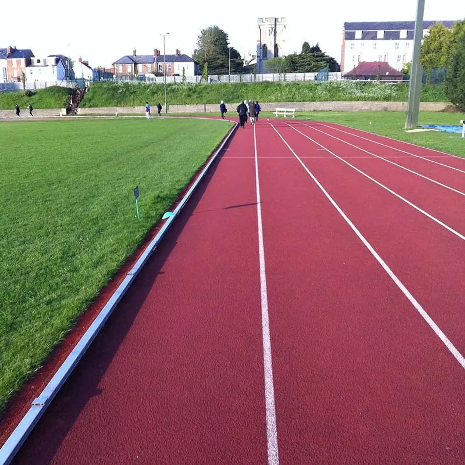 A red, four-lane running track beside a grassy field under a clear blue sky. In the background, a few individuals stroll along the track and buildings are visible. A small sign is placed on the grass near the track.