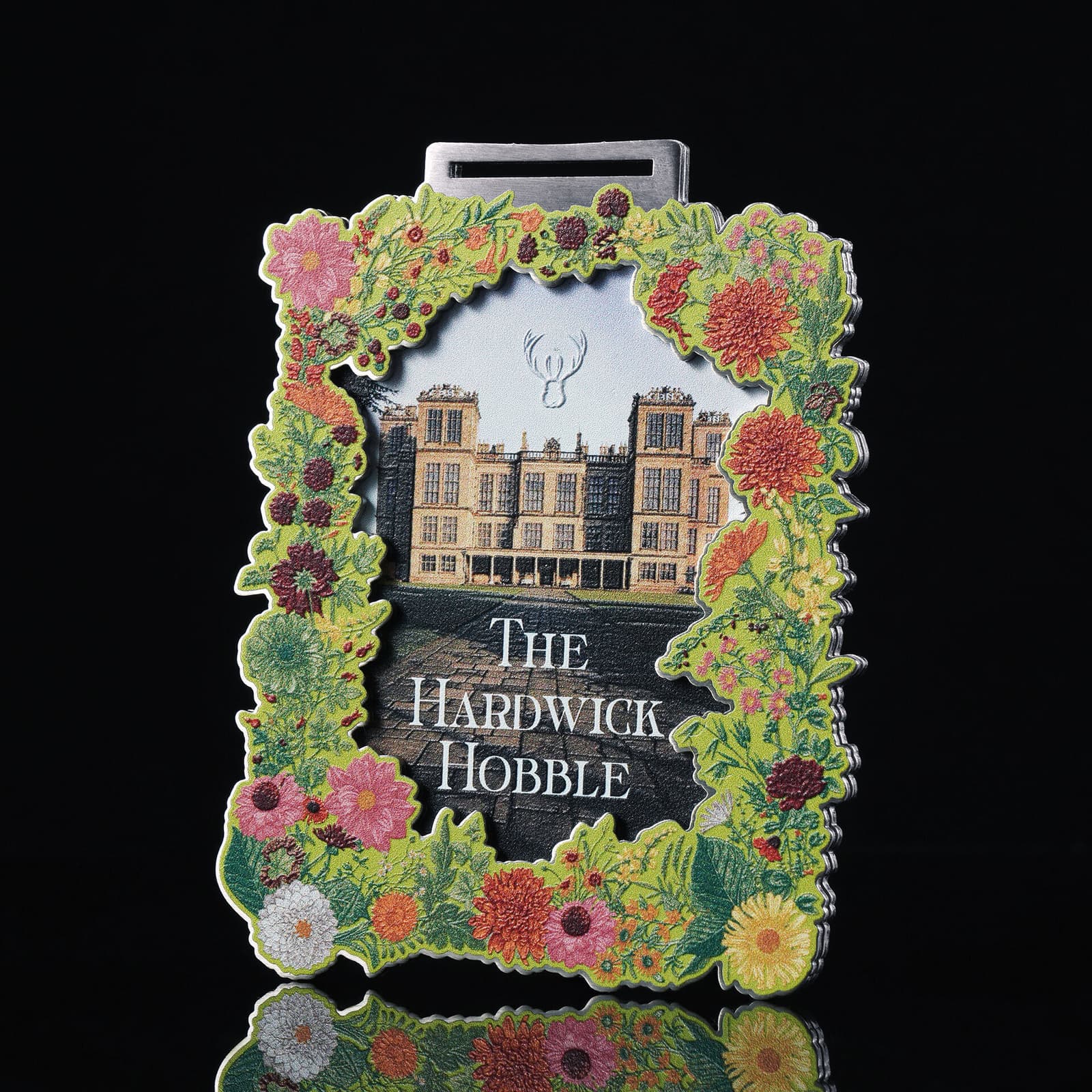 An ornate medal shaped like a flower-laden frame, showcasing an image of a large, historic building. Above the building is a small stag emblem, and below it reads "The Hardwick Hobble." The edges are decorated with colorful flowers and greenery.