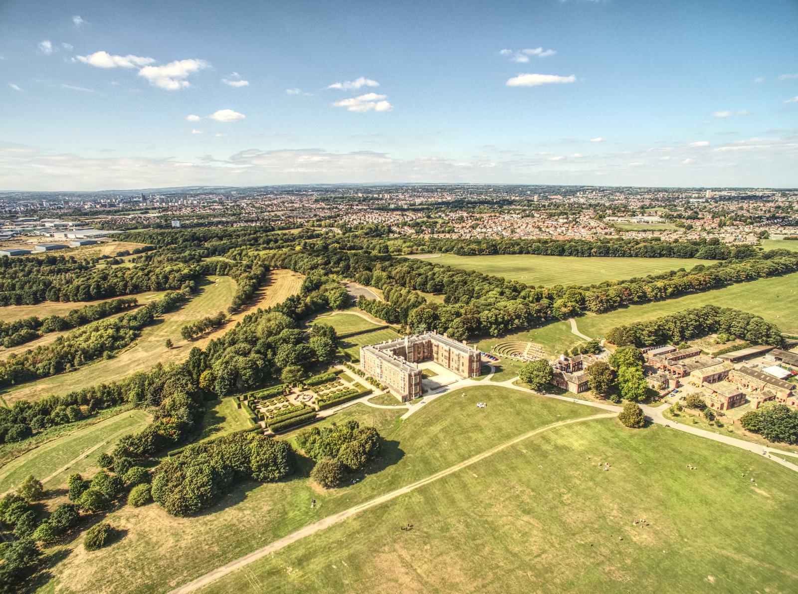Aerial view of a large mansion surrounded by expansive green fields and trees under a clear blue sky. A cityscape is visible in the background, with scattered clouds enhancing the picturesque scenery.