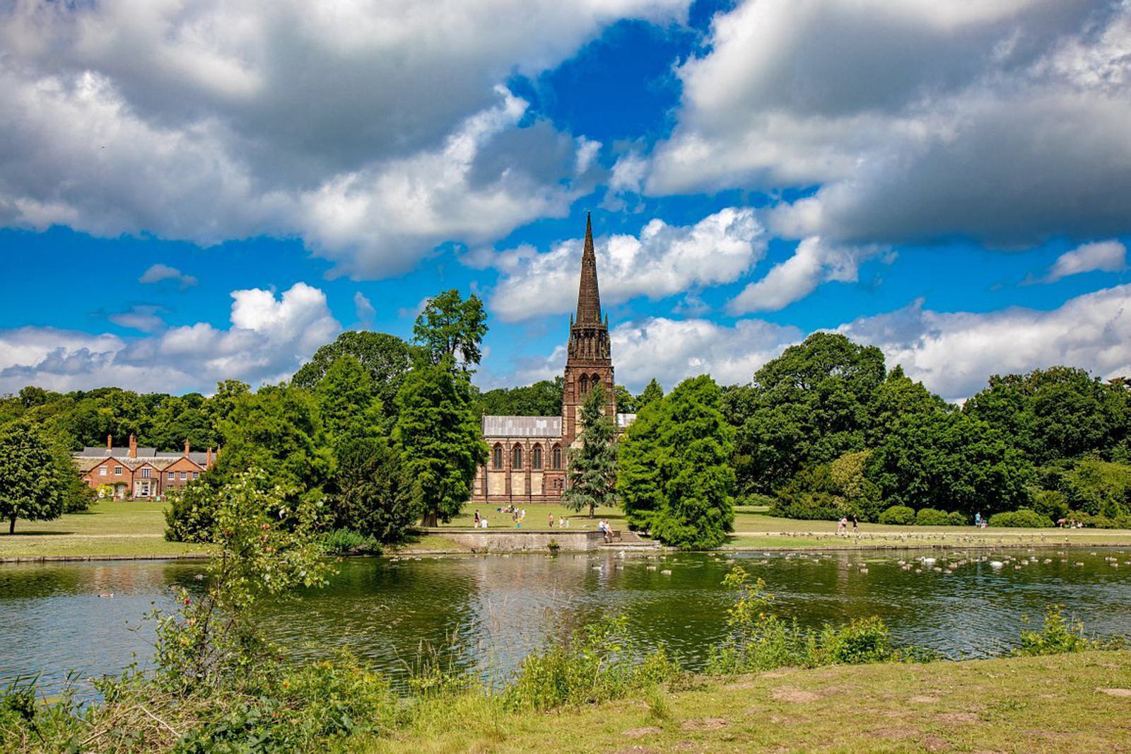 A picturesque scene of a church with a tall spire surrounded by trees near a tranquil lake on a sunny day. The sky is partly cloudy, and the area is lush and green, reflecting a peaceful atmosphere.