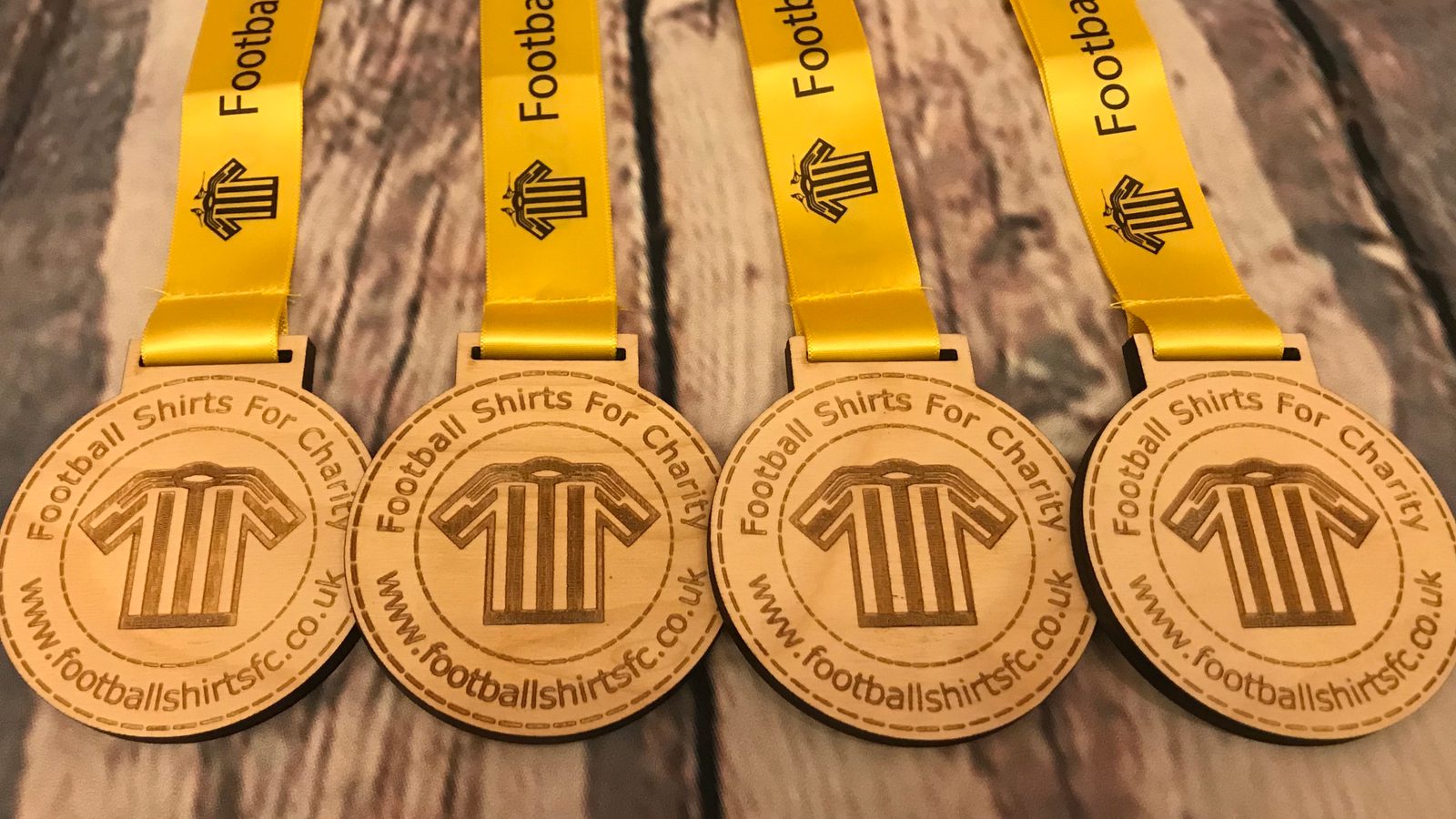 Four wooden medals with yellow ribbons, each engraved with a football shirt icon and the text "Football Shirts for Charity" and the website "www.footballshirtsfc.co.uk," are displayed on a wooden surface.