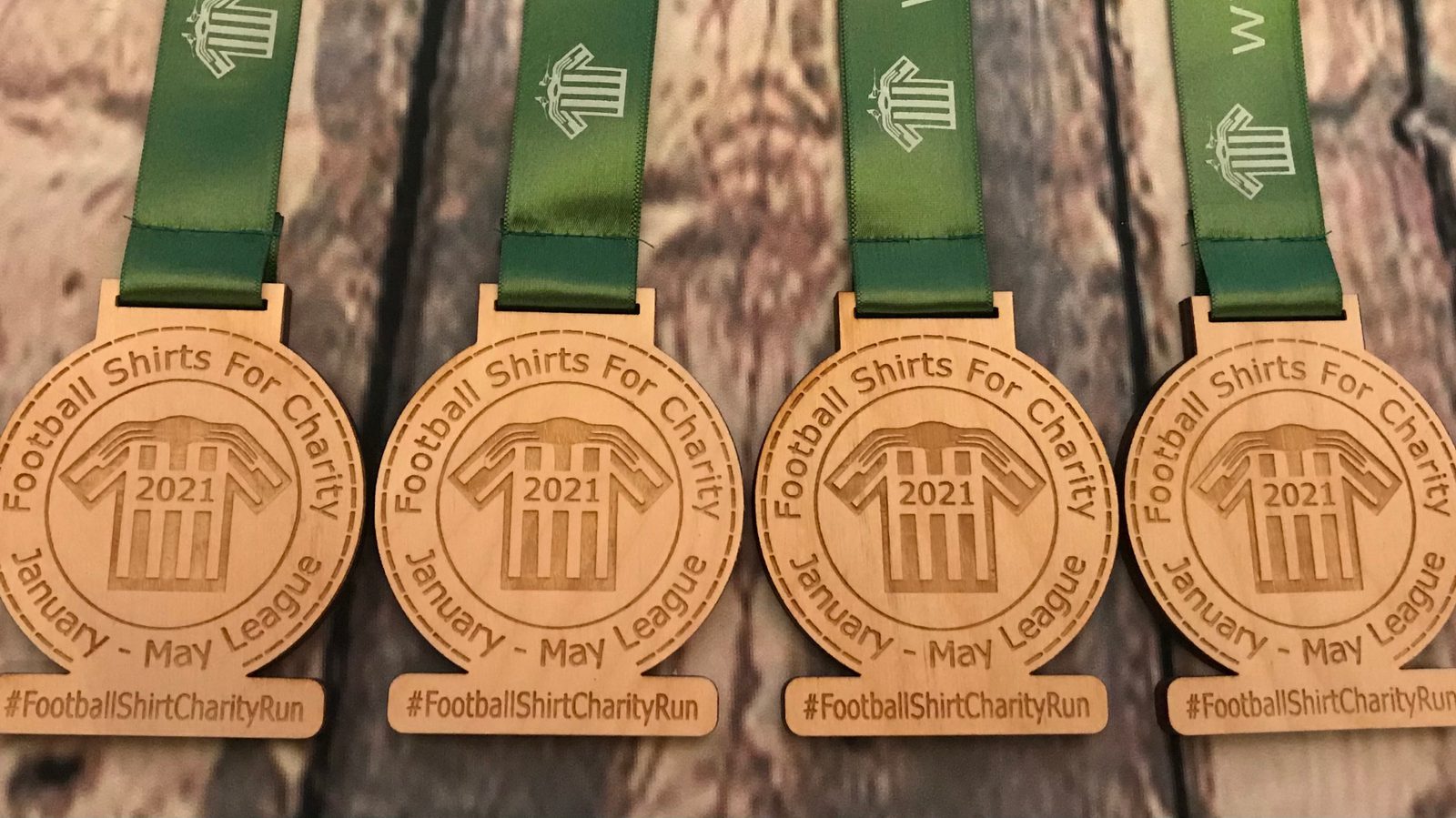 Four wooden medals with green ribbons are lined up. Each medal features the text "Football Shirts For Charity" and "January - May League 2021" with a football jersey icon in the center. Below the medals, the hashtag "#FootballShirtCharityRun" is visible.