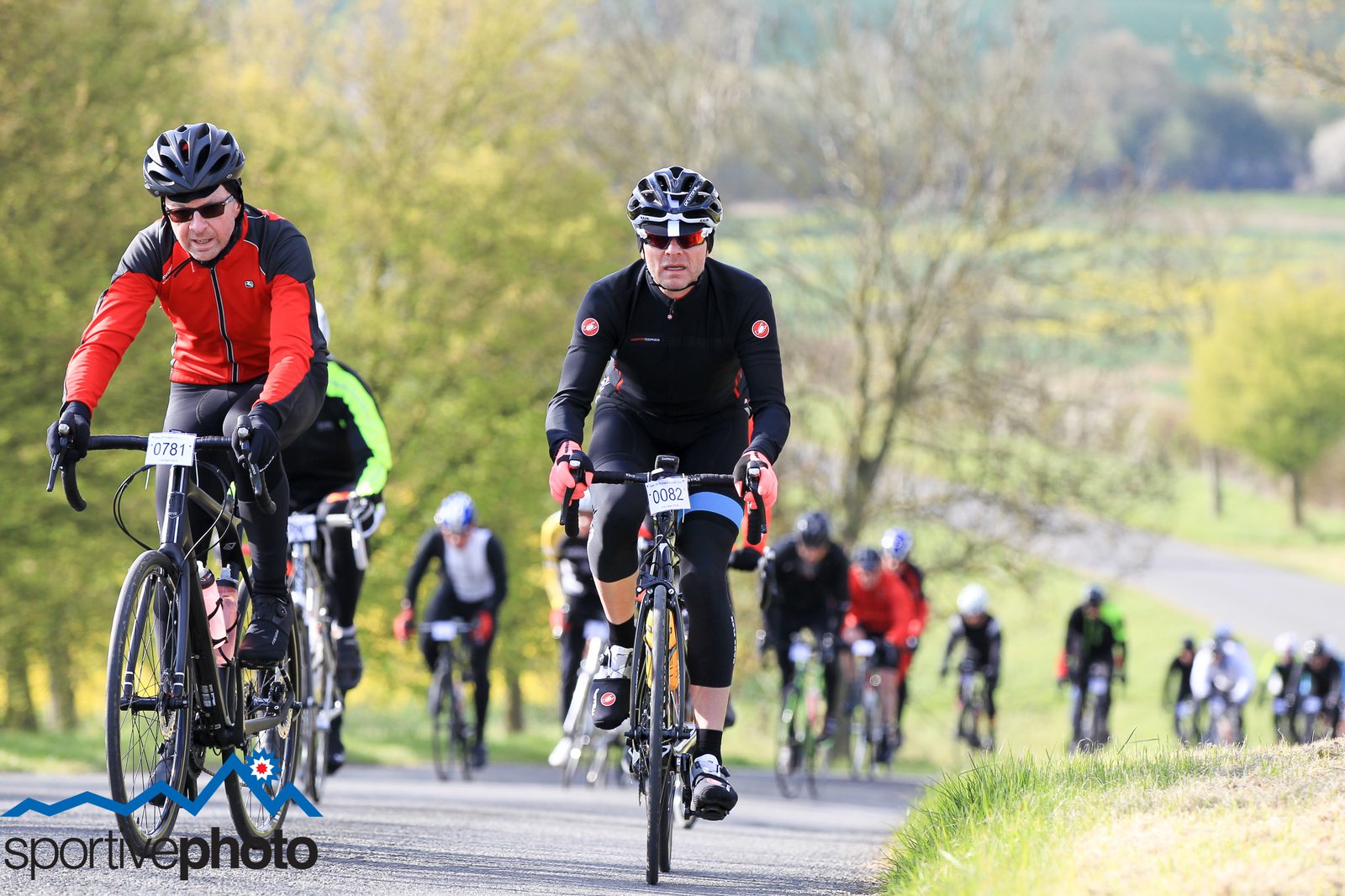 Cyclists participate in a road cycling event on an open countryside road. The riders, dressed in various cycling gear, pedal uphill in a scenic area with greenery and trees in the background. The sky is clear, and the atmosphere appears energetic and focused.