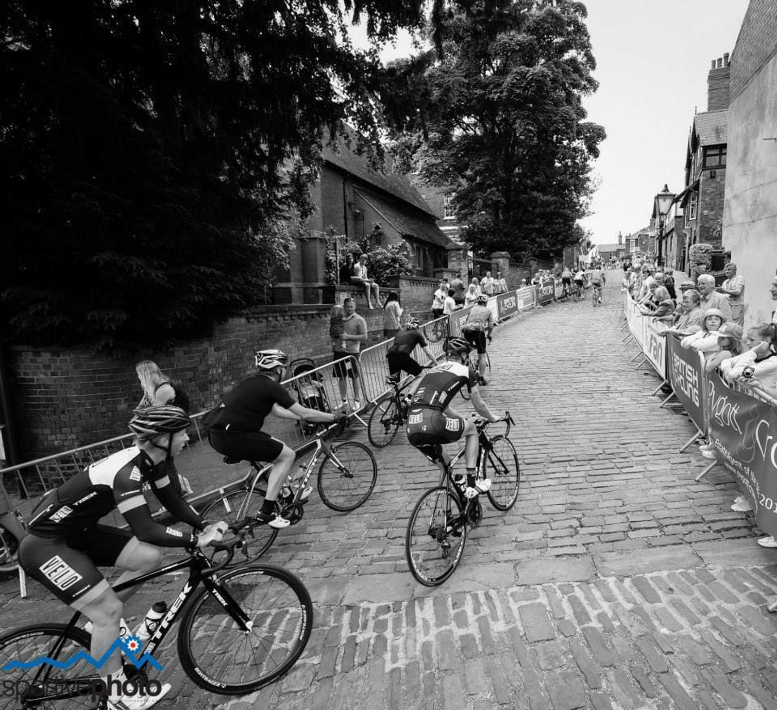 Black and white photo of cyclists during a race navigating a cobblestone street turn. Spectators stand behind barricades, cheering them on. The backdrop consists of historic buildings and tall trees under a partly cloudy sky.