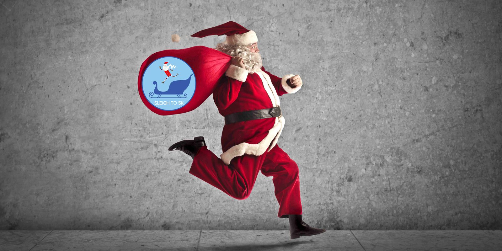 Santa Claus is mid-stride, running with a large red sack slung over his shoulder. The sack is decorated with a logo featuring a blue whale and the text "Submit your wish right to SG." The background is a plain gray wall and floor.