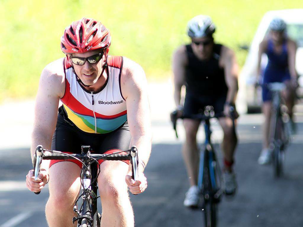 A cyclist wearing a red helmet and sunglasses, riding in front of two other cyclists. The lead cyclist is in a white, red, and rainbow-colored jersey with "Bloodwise" written on it. The background is a sunny road with greenery and a parked car.