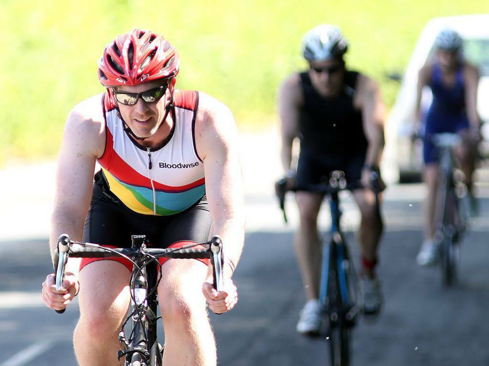 A cyclist wearing a red helmet and sunglasses, riding in front of two other cyclists. The lead cyclist is in a white, red, and rainbow-colored jersey with "Bloodwise" written on it. The background is a sunny road with greenery and a parked car.