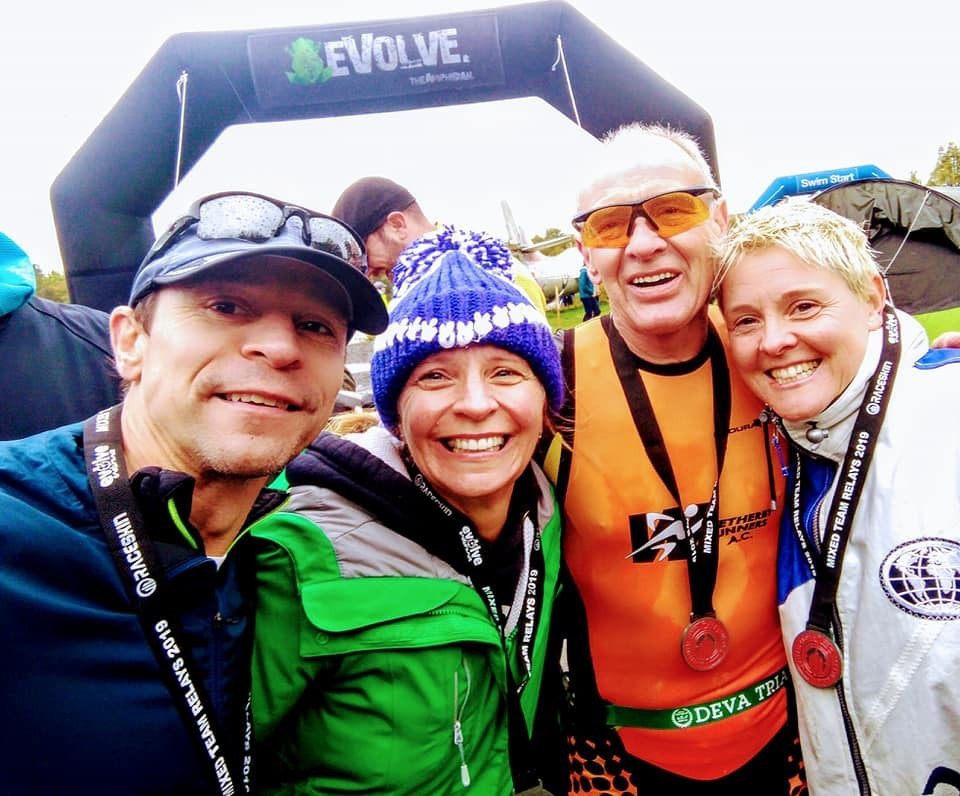 Four smiling people in outdoor gear pose for a selfie at a finish line. They are wearing medals around their necks, indicating they likely participated in a race or endurance event. An arch with the word "EVOLVE" is visible in the background.