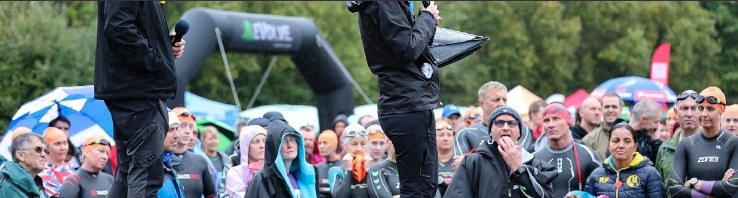 A group of people, mostly dressed in wet suits and swim caps, gather outdoors. Two individuals in the foreground, dressed in black jackets, appear to be addressing the crowd. An inflatable arch and trees are visible in the background, and some attendees hold umbrellas.