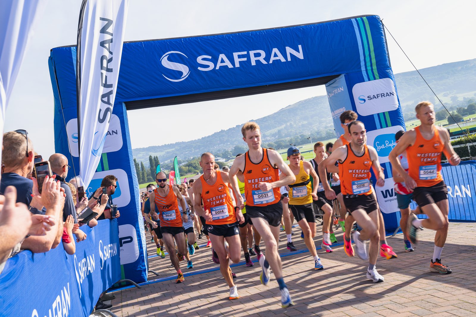 Runners in orange and black jerseys begin a race under a blue archway with the Safran logo. Spectators line the sides, cheering. The background features rolling hills and a clear sky. Banners and flags with "Safran" branding are visible.