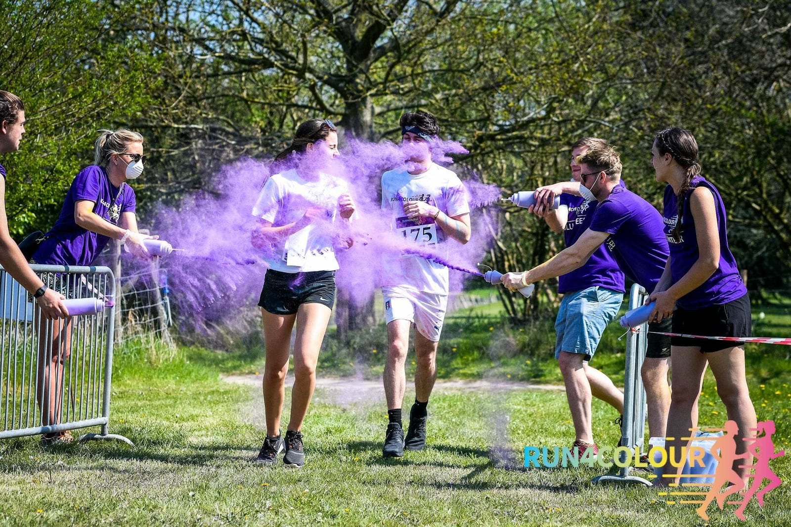 Participants in a color run event are being sprayed with purple powder as they run through a designated area. Two individuals on each side of the path, wearing purple shirts, are throwing the color powder. Other runners and trees are visible in the background.