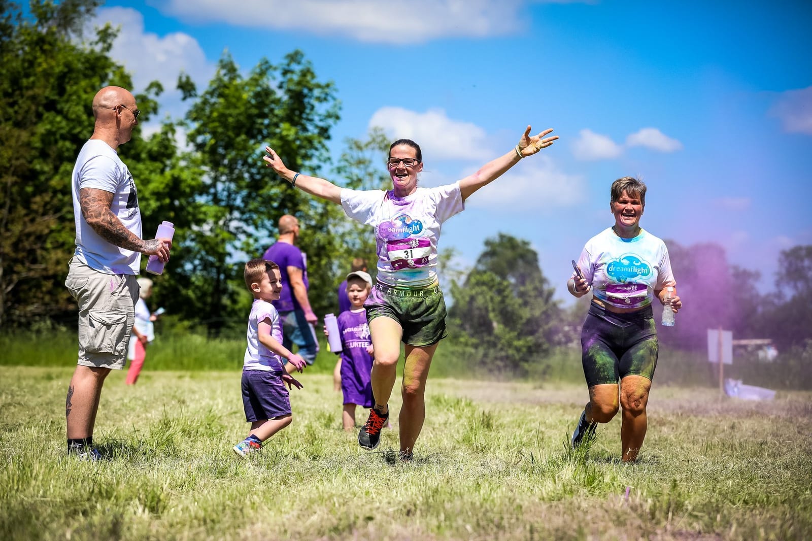 Participants in a Color Run event are joyfully running through a grassy field, surrounded by colorful powder. Two women with arms raised in excitement lead the way, while children and other adults watch and celebrate. The sky is clear with a few scattered clouds.