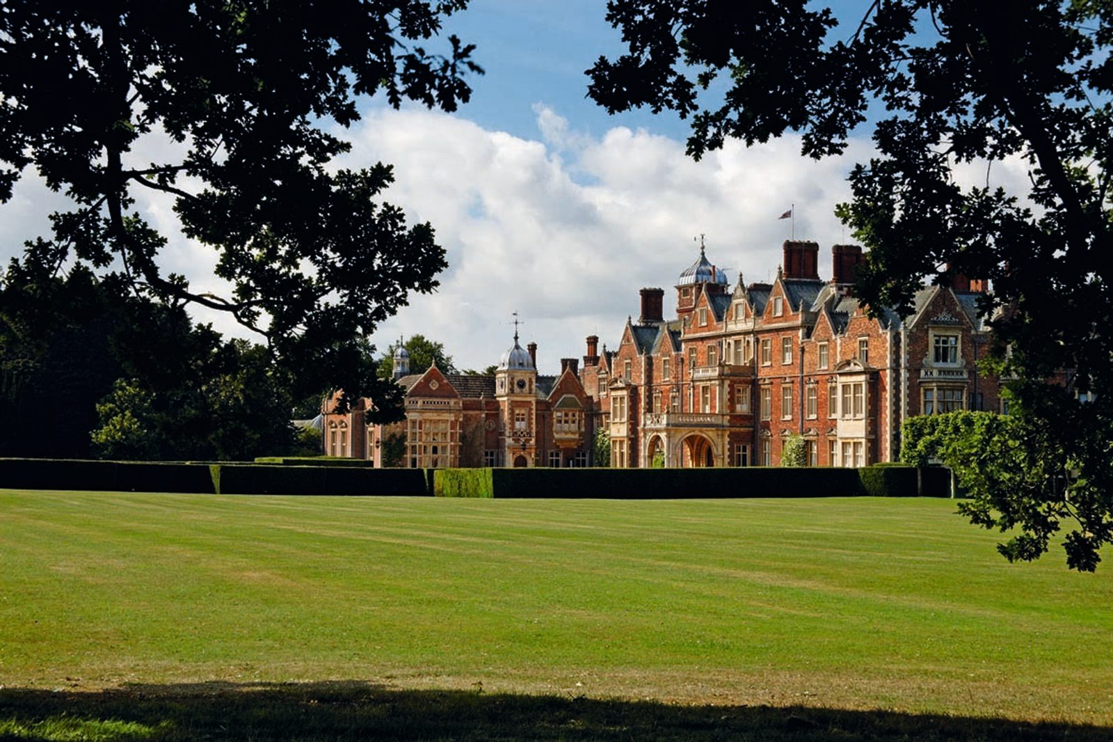 A grand, historic building with intricate brickwork sits amid lush green lawns and manicured hedges, framed by trees. The bright blue sky with scattered clouds serves as a backdrop, highlighting the regal architecture and peaceful surroundings.