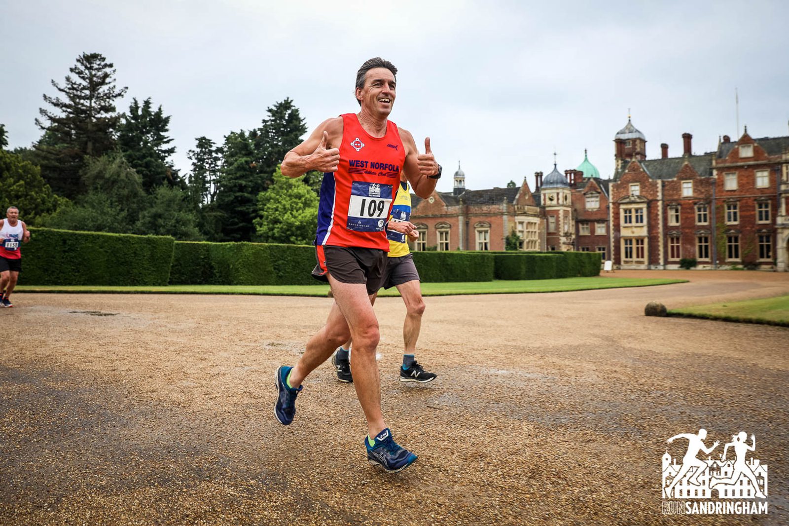 A runner in athletic gear smiles and gives thumbs-up while participating in a race near an old ornate building and well-kept hedges. Another runner is visible in the background, slightly obscured. The ground is wet, and the sky is overcast. There is a "Sandringham" logo in the corner.