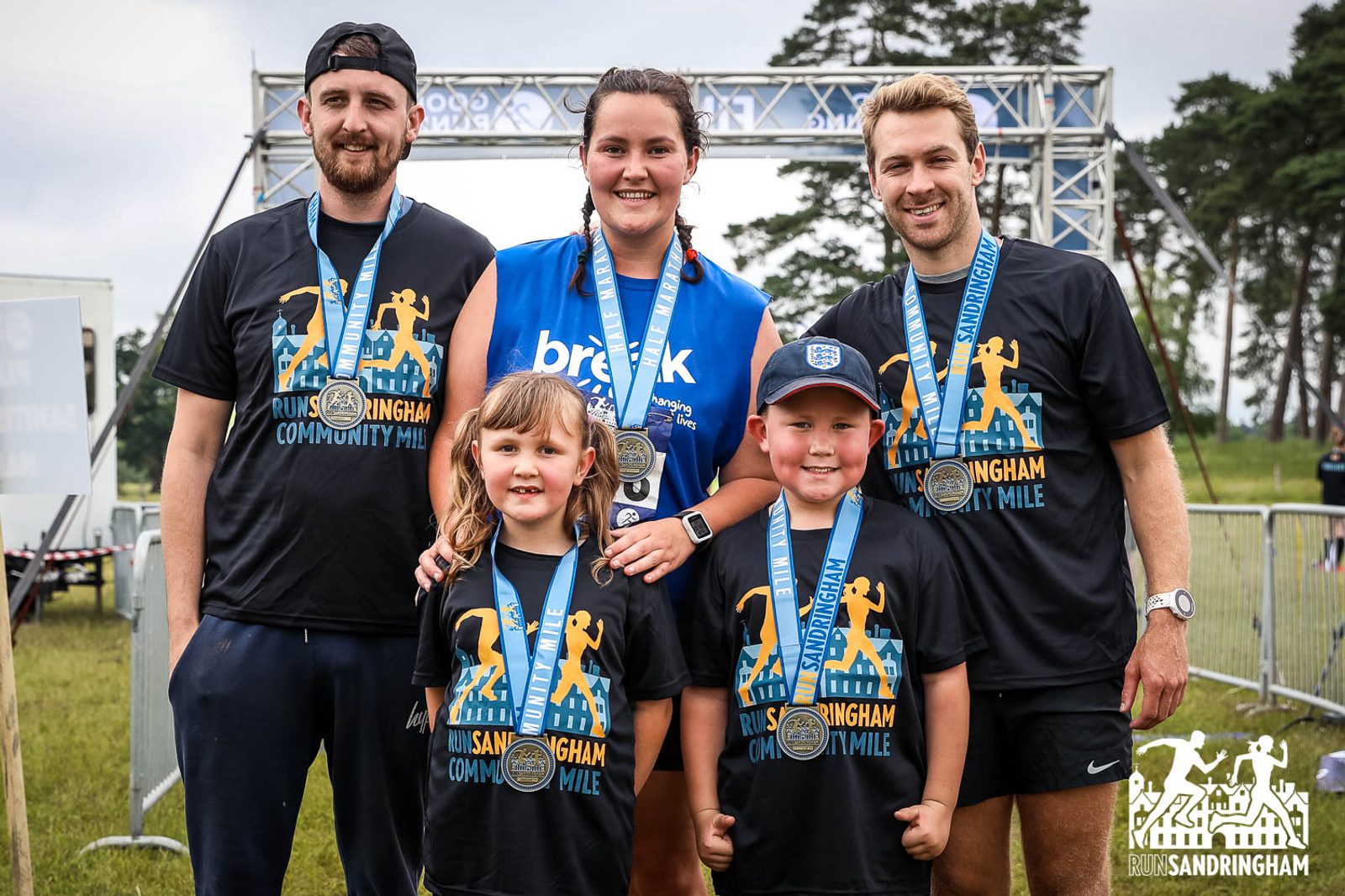 A group of five individuals stands smiling with medals around their necks in front of a finish line arch. The adults are wearing black "Run Sandringham Community Mile" shirts, and the child in the center sports a blue shirt. Trees and greenery are visible in the background.