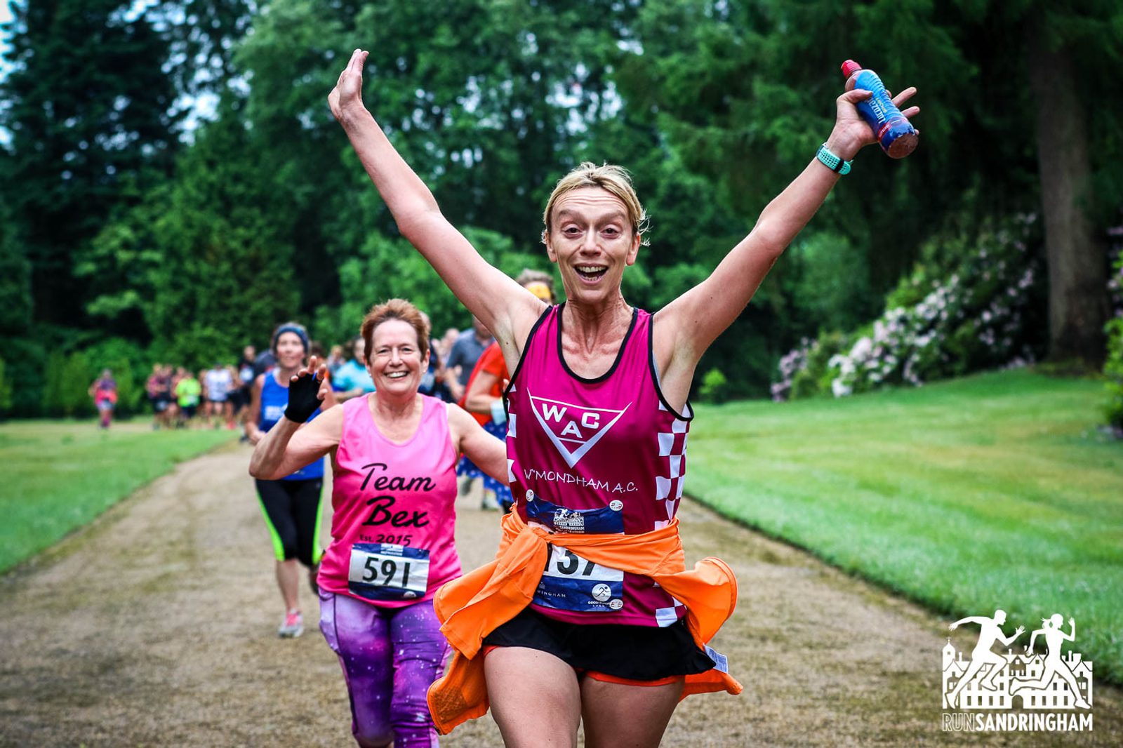 A woman in a pink tank top and orange jacket tied around her waist runs with both arms raised in celebration, holding a water bottle. Behind her, other runners, including one in a pink "Team Bex" shirt, are also running. The background features green trees and grass.