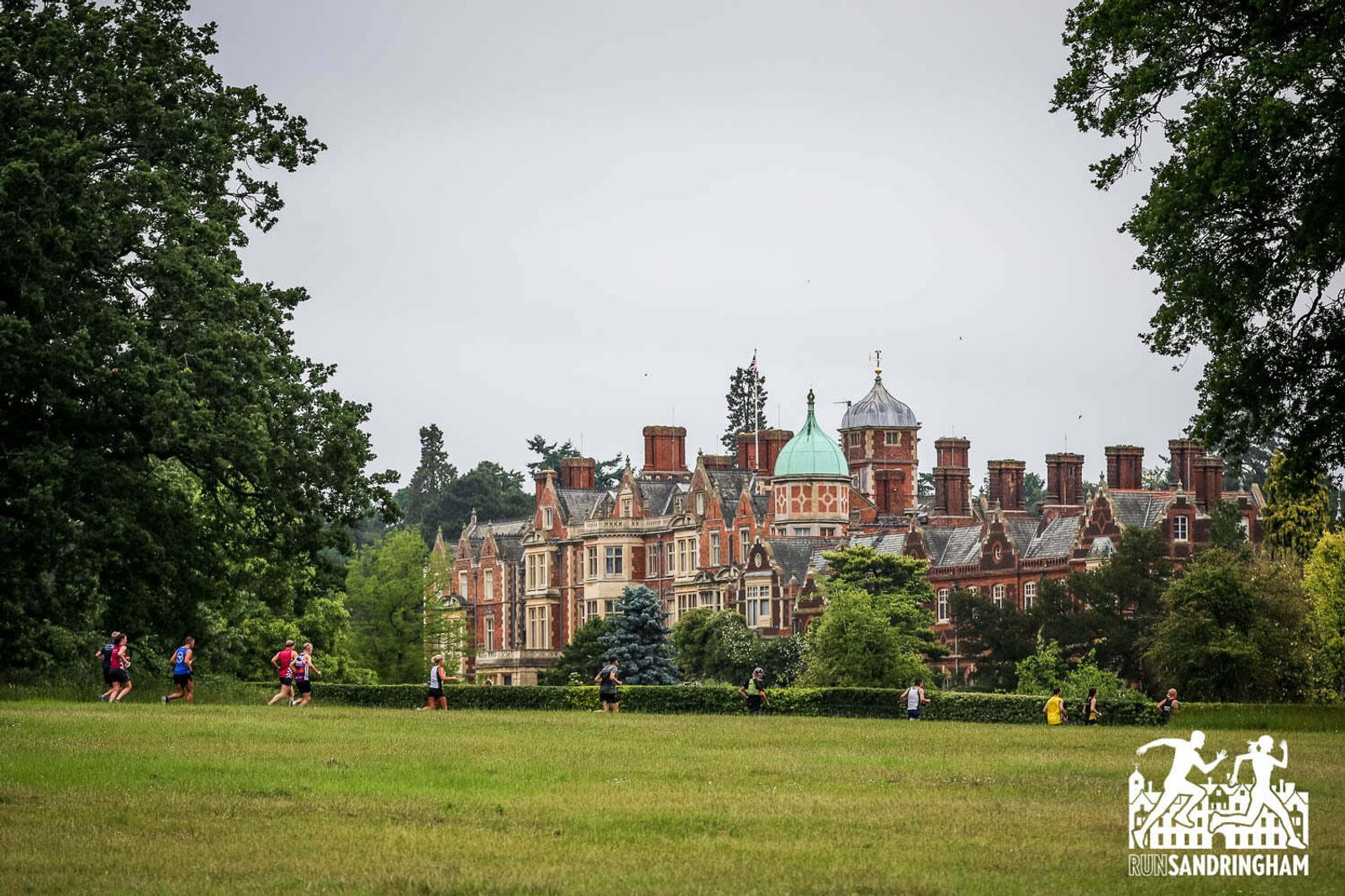 Runners jog along a grassy field with the Sandringham Estate, a large, ornate mansion with a mix of architectural styles and chimneys, in the background. Trees surround the estate, and the sky is overcast.