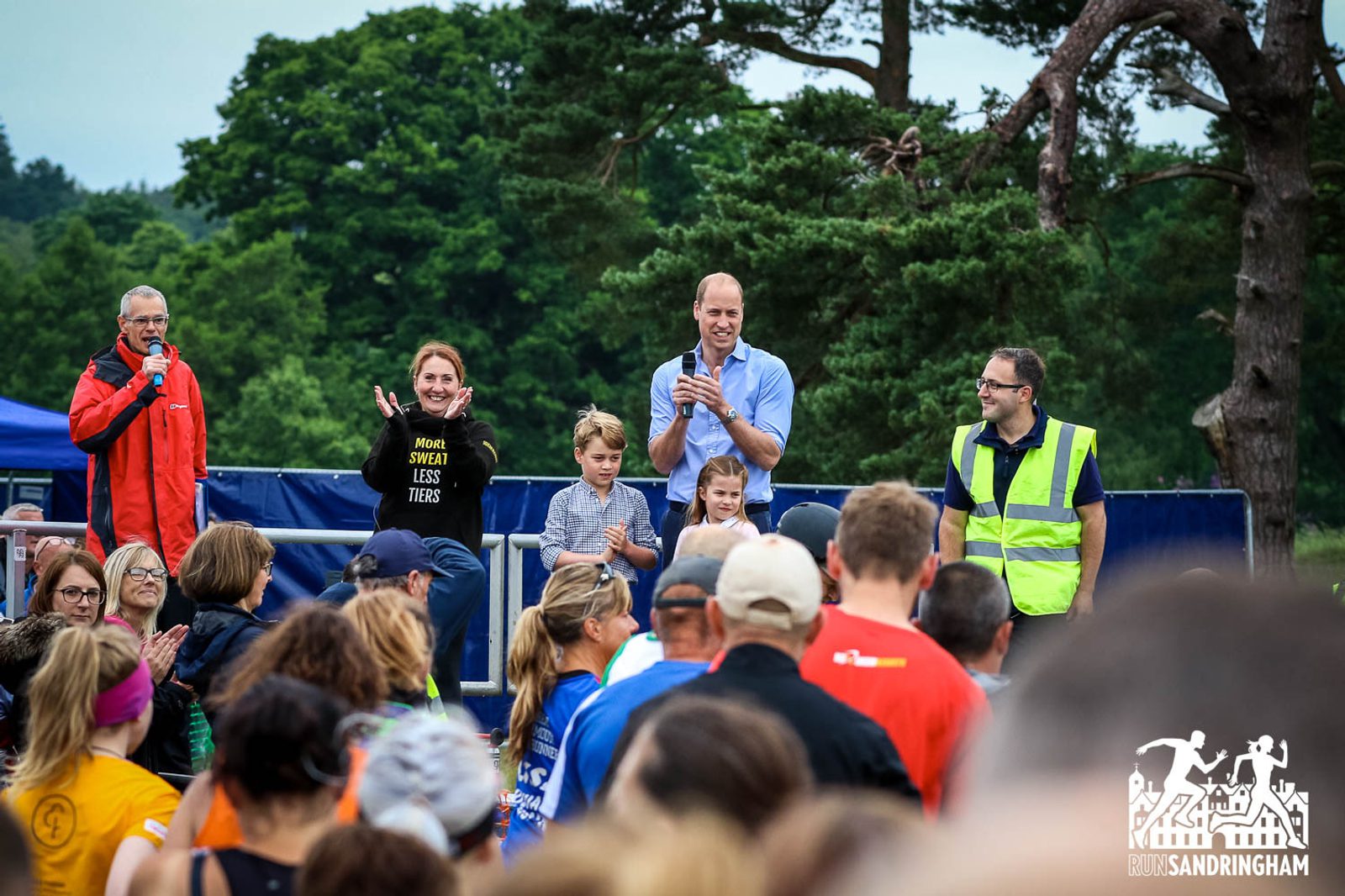 A crowd of runners gathers at an outdoor event. A man in a high-visibility vest, a woman with a microphone, and a man holding two children stand on a stage applauding. Dense greenery and trees are visible in the background.