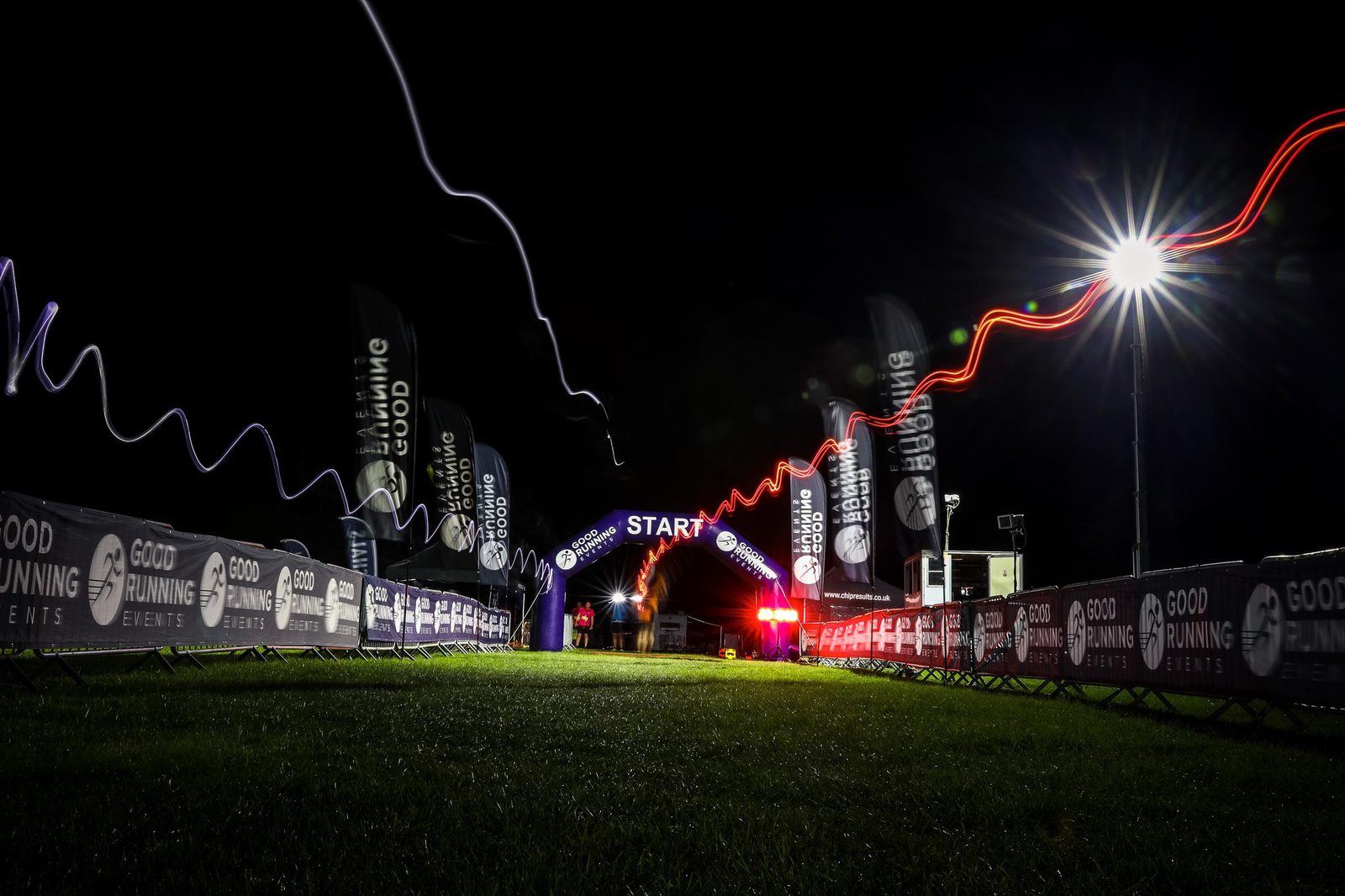 Nighttime view of a race starting line. The banner reads "START," illuminated by spotlights and surrounded by dynamic light trails. Two parallel fences display "GOOD RUNNING EVENTS" banners, guiding participants to the starting point on a grass field.