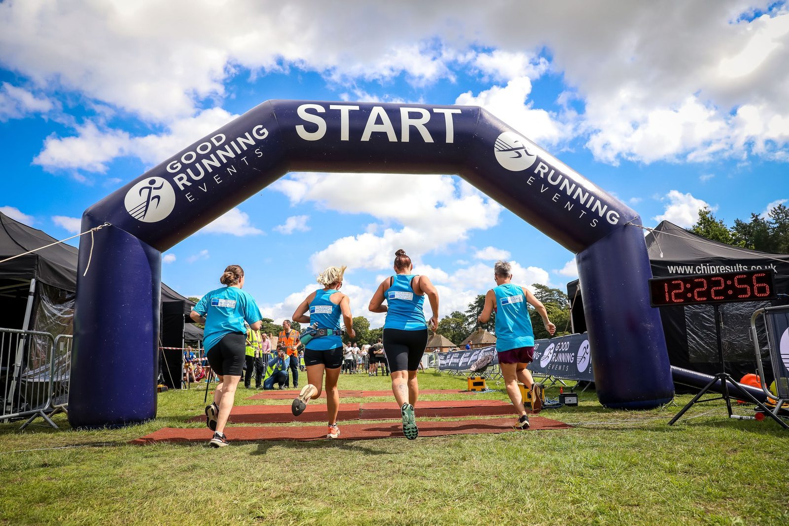 Four runners in blue attire are passing under a purple inflatable start arch with "Good Running Events" printed on it. The timing clock to the right shows 12:22:56. The sky is partly cloudy, and there are spectators and tents in the background.