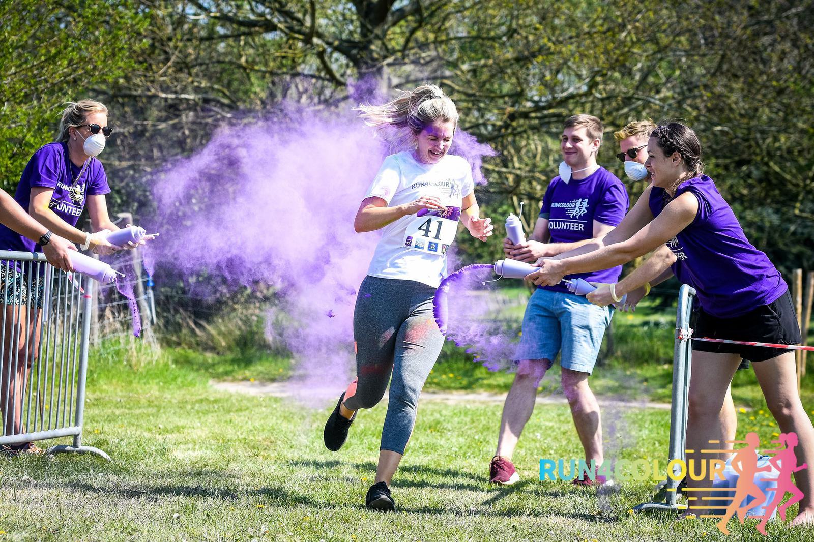 A runner wearing a white t-shirt and gray pants is sprayed with purple powder by volunteers as she runs in a vibrant outdoor color run. Other participants and spectators can be seen around her, with trees and green grass in the background.