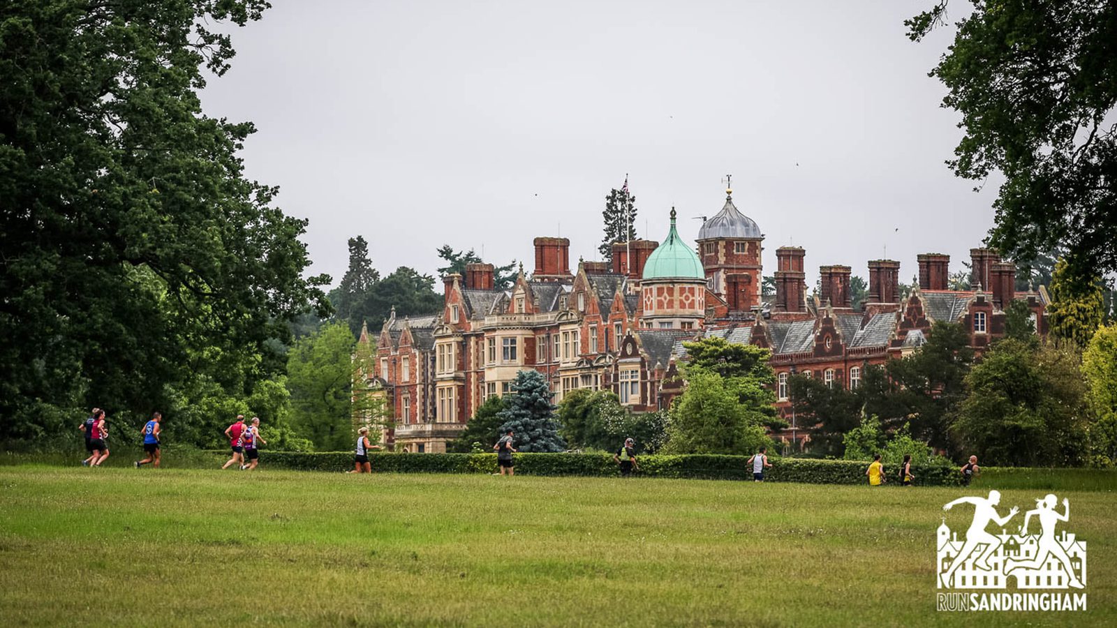 A group of people in athletic clothing run across a grassy field with a large, historic, red-brick building and lush greenery in the background. The event logo "Run Sandringham" is visible in the bottom right corner.