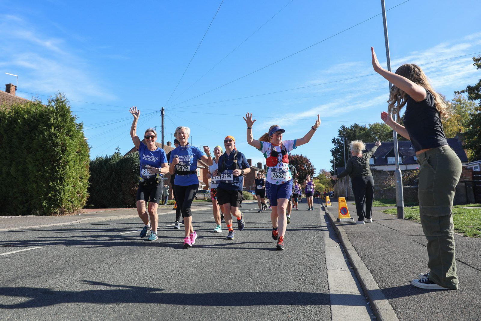 A group of marathon runners in race bibs joyfully raise their arms as they run down a sunny street. A cheering spectator on the right waves at them. Trees, houses, and blue sky are visible in the background.
