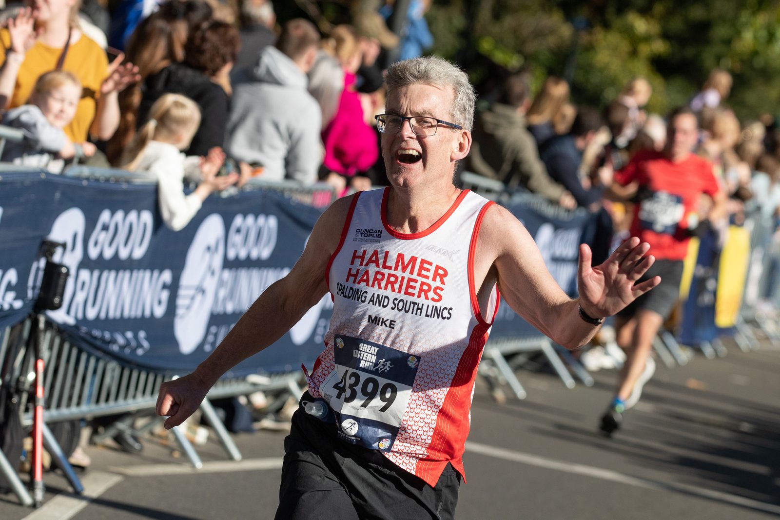 A man with gray hair and glasses is running in a race. He is wearing a red and white "Halmer Harriers" running vest and a race bib numbered 4399. He looks happy and energetic. Spectators are cheering behind barriers, and other runners are visible in the background.