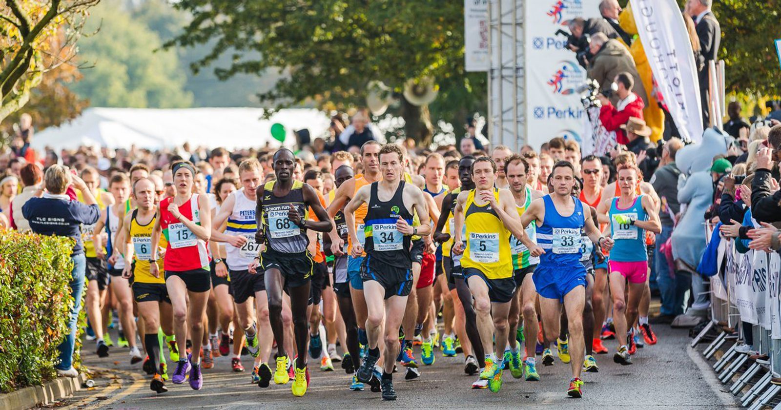 A large group of runners, wearing race bibs and colorful athletic gear, starts a marathon on a tree-lined street. Spectators stand on both sides of the street, cheering and taking photos as the runners pass by.
