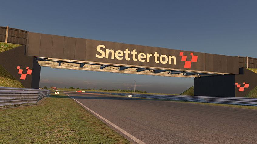 A view of the Snetterton race circuit, showing a track under a bridge with "Snetterton" and a red checkered flag logo displayed on the bridge. The track is surrounded by grass with a clear sky in the background.